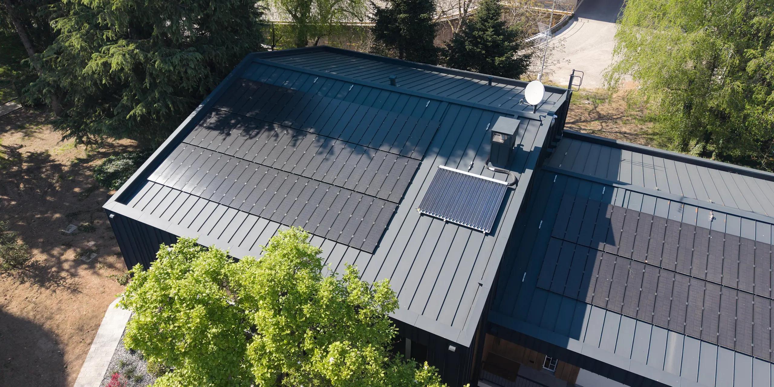 Luftaufnahme der Villa Castellanza mit PREFALZ Dach in P.10 Anthrazit und harmonisch integrierten PREFALZ Solarmodulen. Die klaren Stehfalzlinien prägen die moderne Architektur und unterstreichen die hochwertige PREFA Gebäudehülle. Umgeben von altem Baumbestand vereint das Objekt nachhaltige Energiegewinnung, langlebige Aluminiumqualität und zeitlose Ästhetik auf elegante Weise.