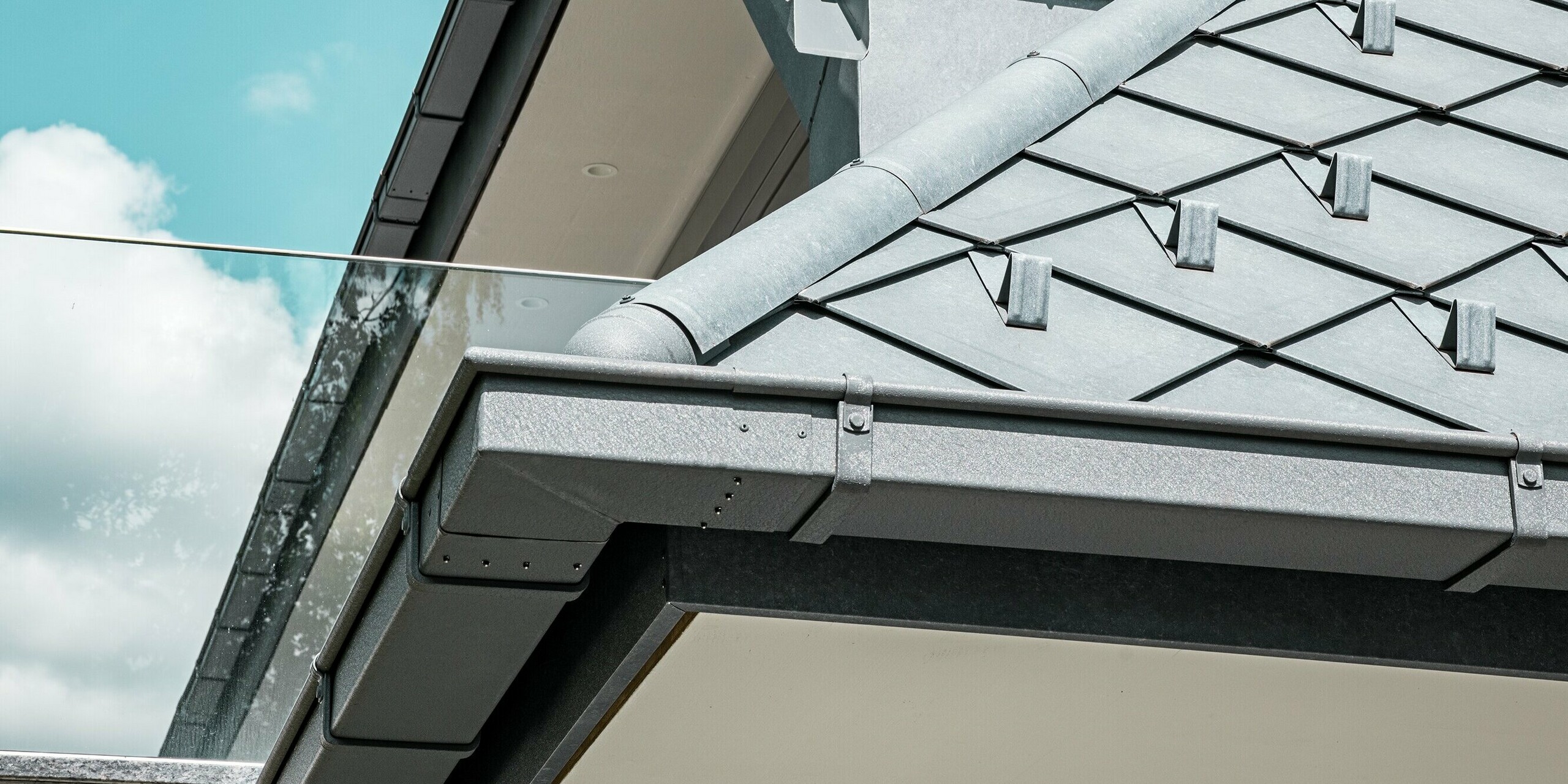 Nahaufnahme des Daches eines modernen Mehrfamilienhauses mit PREFA Dachrauten 29 × 29 in P.10 Steingrau. Die Dachkante ist elegant mit einer formschönen Kastenrinne und einem Ablaufrohr aus Aluminium ausgestattet, die für eine effiziente Dachentwässerung sorgen. Der Balkon mit Glaswand ergänzt das minimalistische Design des Hauses und setzt einen Kontrast zur robusten Dachkonstruktion. Die hochwertigen Materialien und präzise Verarbeitung bieten sowohl ästhetische als auch funktionale Vorteile, die das moderne Wohnambiente unterstreichen.