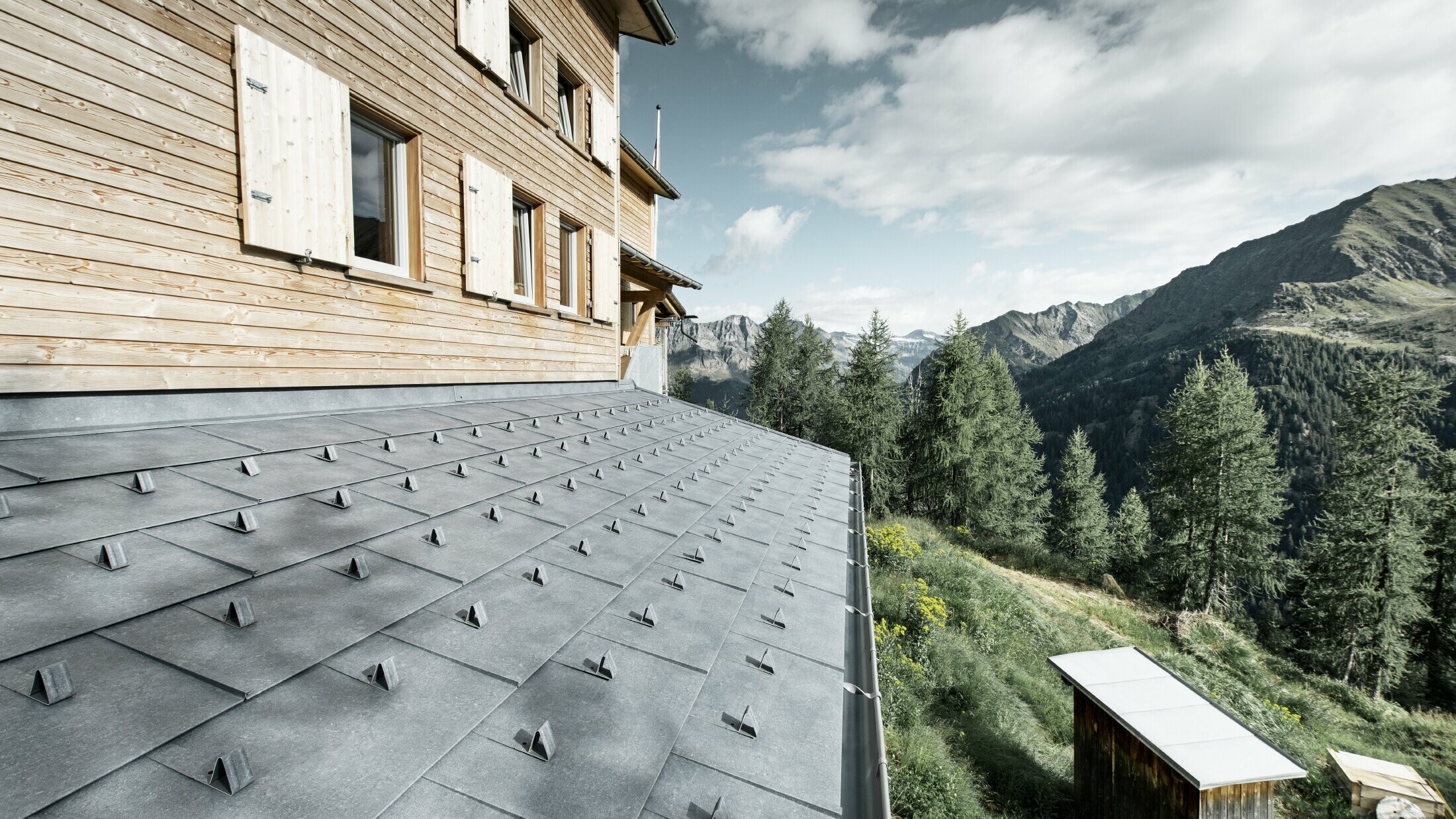 Particolare di una tettoia su una capanna di legno. La tettoia è rivestita con pannelli di alluminio PREFA FX.12 in grigio pietra con naso fermaneve. La grondaia è tenuta in posizione con dei tiranti.