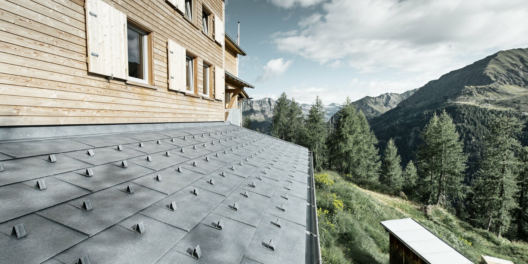 Particolare di una tettoia su una capanna di legno. La tettoia è rivestita con pannelli di alluminio PREFA FX.12 in grigio pietra con naso fermaneve. La grondaia è tenuta in posizione con dei tiranti.