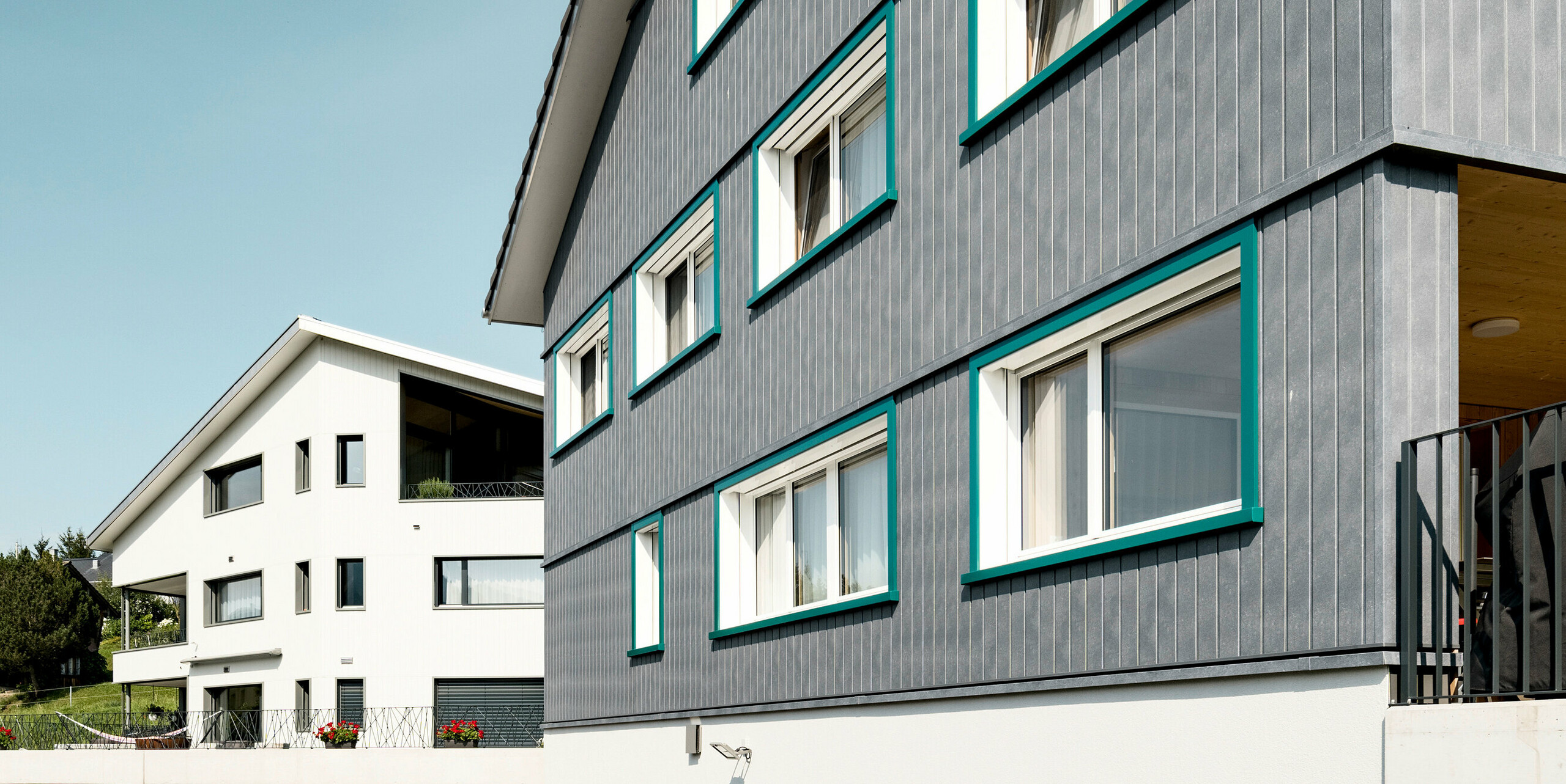 Ansicht zweier benachbarter Mehrfamilienhäuser in Weissbad, Schweiz, wobei das vordere Gebäude mit seiner PREFA Siding Fassade in P.10 Steingrau einen starken visuellen Eindruck vermittelt. Die vertikal montierten Sidings schaffen eine elegante und schützende Hülle, während die türkisen Fensterrahmen farbliche Akzente setzen. Das Bild fängt das Zusammenspiel der modernen Materialien mit der traditionellen Architektur ein und demonstriert die vielseitige Einsetzbarkeit von PREFA Aluminiumprodukten.