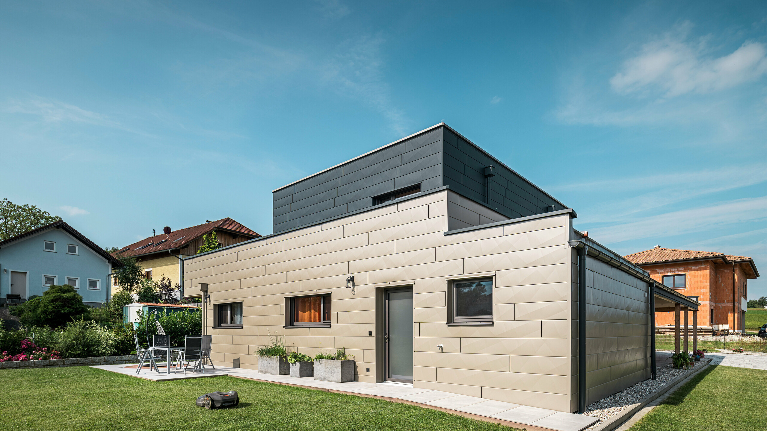 Modernes Einfamilienhaus in Wippenham, Österreich, mit zweifarbiger PREFA Aluminiumfassade in Bronze und Anthrazit. Die Fassade ist mit Siding.X Fassadenpaneelen gestaltet, die für eine elegante und zeitgemäße Optik sorgen. Die integrierte PREFA Dachentwässerung mit Wasserfangkasten und Ablaufrohr sowie eine gepflegte Gartenanlage mit Terrasse betonen das moderne Design des Hauses.