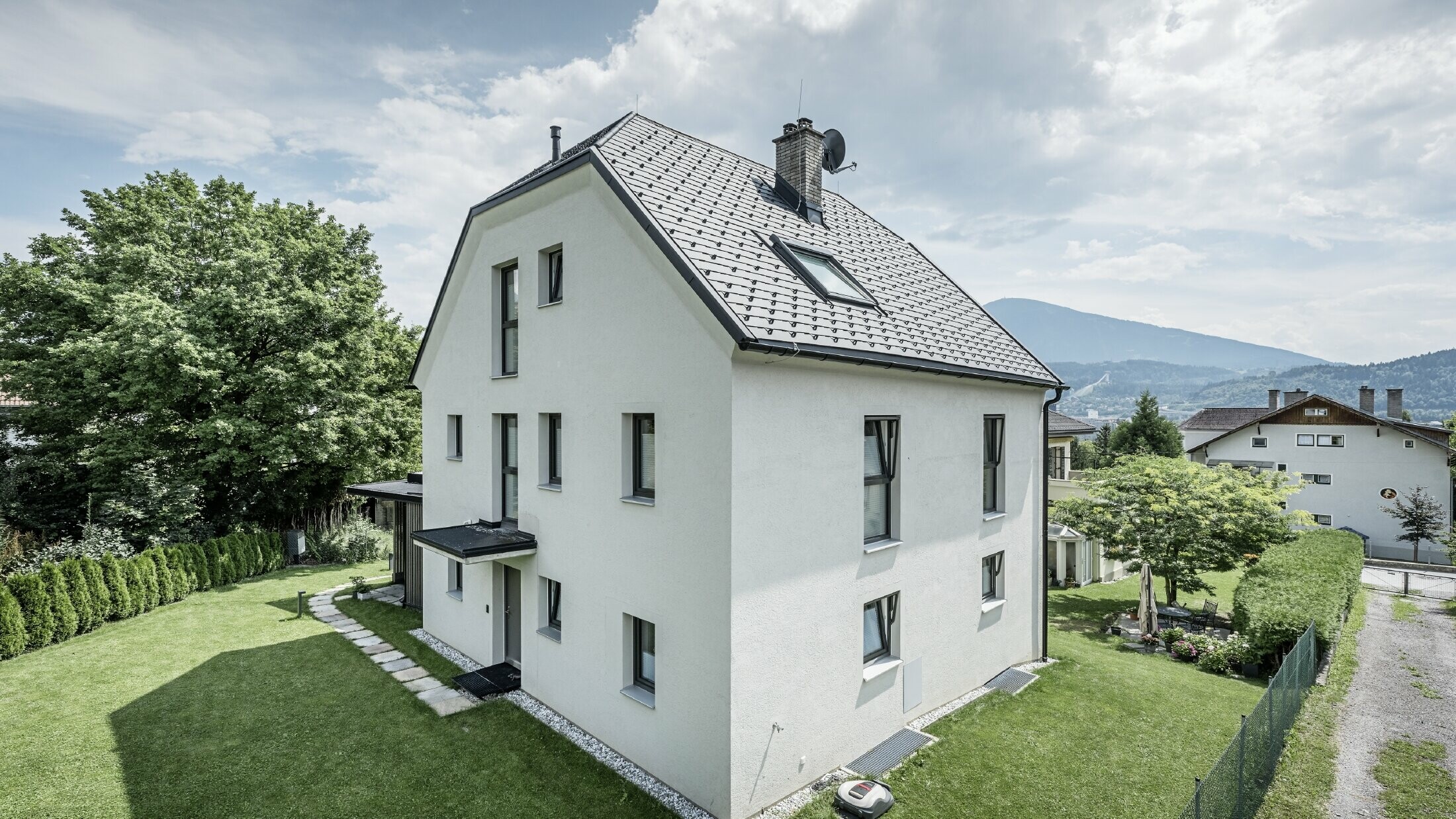 Moderna casa unifamiliare a tre piani con facciata in intonaco chiaro e tetto a due falde in alluminio PREFA grigio scuro. L'edificio presenta diverse finestre strette e un sistema di smaltimento acque piovane in antracite. Si trova in un giardino ben curato con prato, alberi e siepi, sullo sfondo si vedono altre case e un paesaggio montuoso.