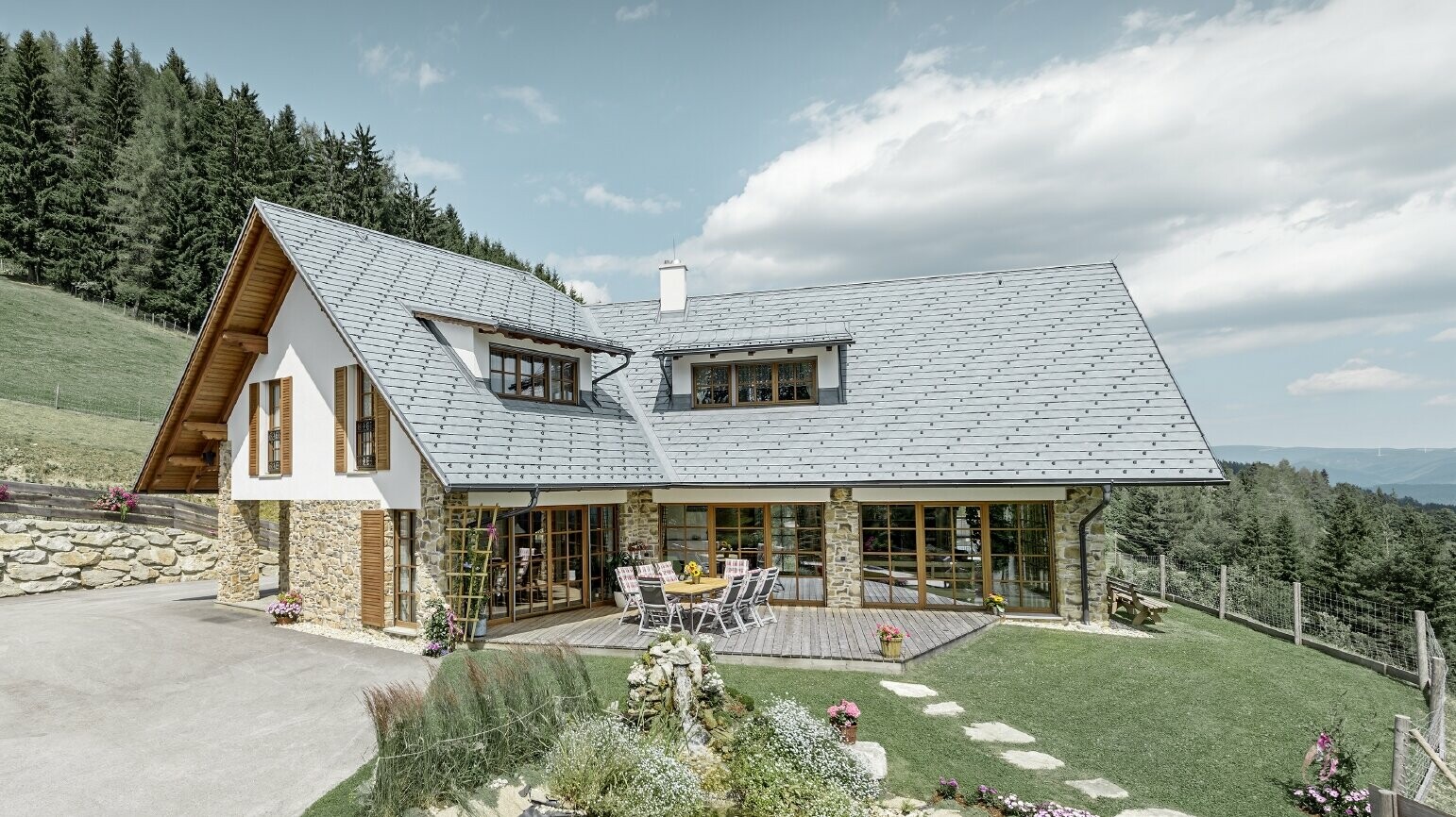 La casa unifamiliare in pendenza, costruita a forma di L, ha un tetto a due falde con due abbaini. Il tetto è stato rivestito con la scandola PREFA in grigio pietra. In primo piano c'è un laghetto e sullo sfondo c'è un bosco.
