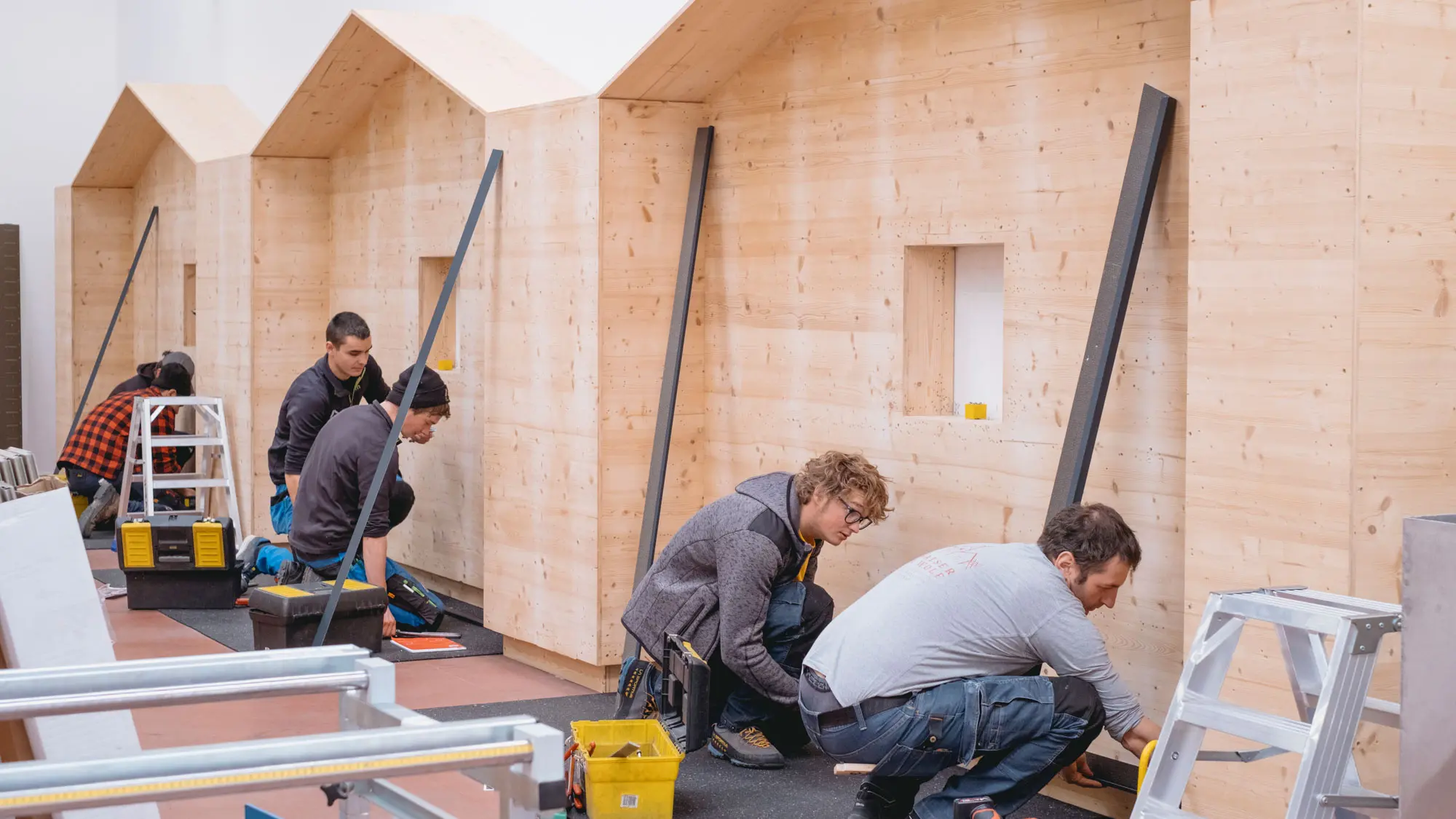 Mehrere Teilnehmer der PREFA Academy in Italien arbeiten an der Montage von Fassadenstrukturen. Sie befestigen Metallprofile an Holzwänden, um die Konstruktion einer Fassade zu üben. Werkzeuge und Materialien sind um sie herum verteilt, während sie sich intensiv auf die praktischen Schulungsaufgaben konzentrieren. Im Hintergrund sind Holzwände in Form von Häusern zu sehen, die als Übungsobjekte dienen.