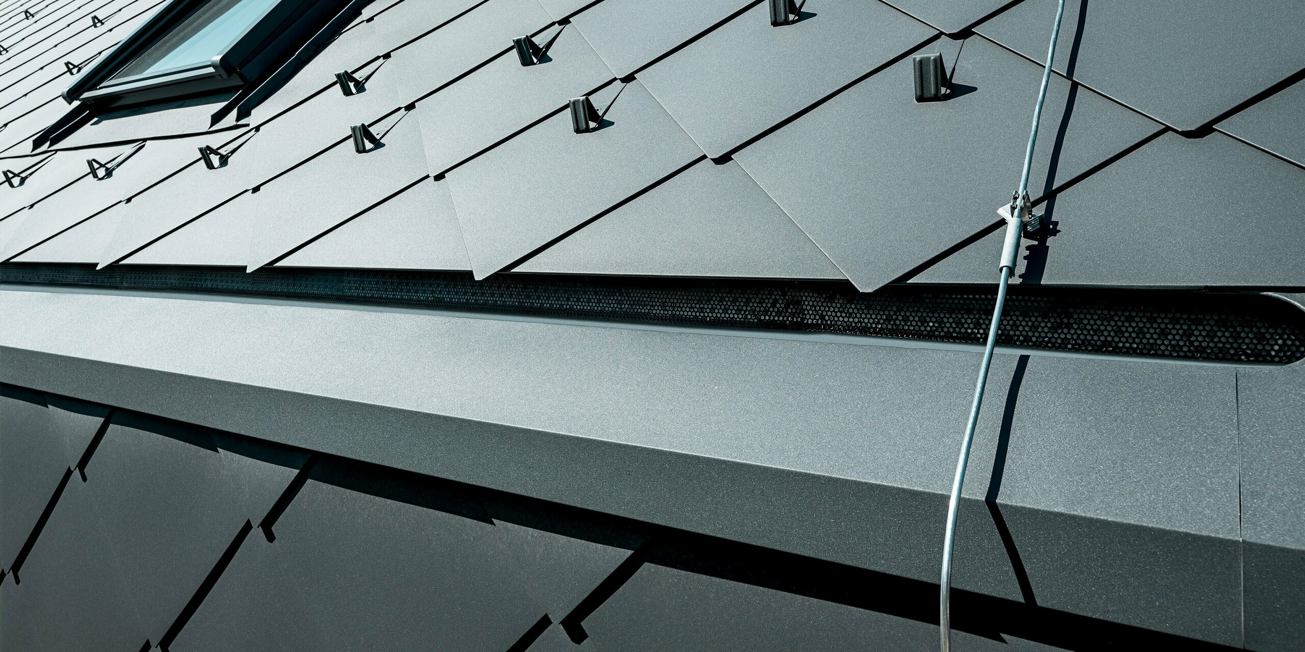 Detailaufnahme des PREFA Dachs mit Dachrauten 44 × 44 in P.10 Dunkelgrau am Einfamilienhaus in Trnava‑Modranka. Das Bild zeigt die präzise verlegte Aluminiumdeckung mit funktionalen Schneestoppern sowie einem integrierten Dachfenster. Die matte Oberfläche der Rauten reflektiert das Licht gleichmäßig und betont die hochwertige Verarbeitung. Die langlebigen PREFA Aluminiumprodukte bieten maximale Stabilität, Witterungsbeständigkeit und eine moderne, technisch anmutende Optik, die dem Gebäude seine markante Architektur verleiht.