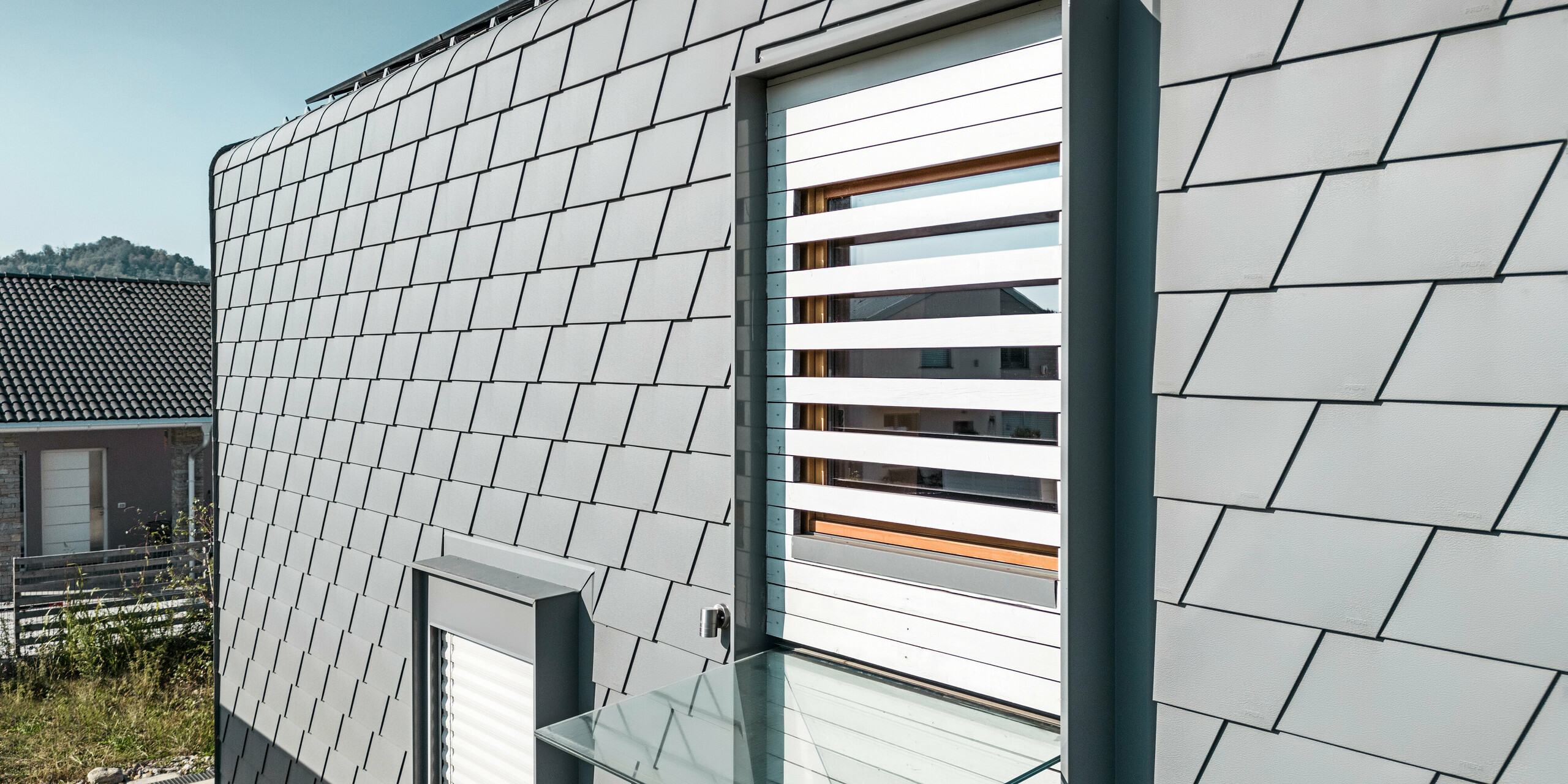 Detailaufnahme eines Fensters mit weißen Außenjalousien. Die Fassade des modernen Hauses ist mit PREFA Wandschindeln in der Farbe P.10 Hellgrau verkleidet. Das Dach ist mit PREFA Dachschindeln in P.10 Hellgrau eingedeckt. Die klare Geometrie der Schindeln ergibt eine strukturierte Oberfläche, die durch die natürlichen Schattenwürfe zusätzlich betont wird. Das Fenster mit den Außenjalousien durchbricht das Muster und fügt ein warmes, natürliches Element in das kühle Grau der Schindeln ein.