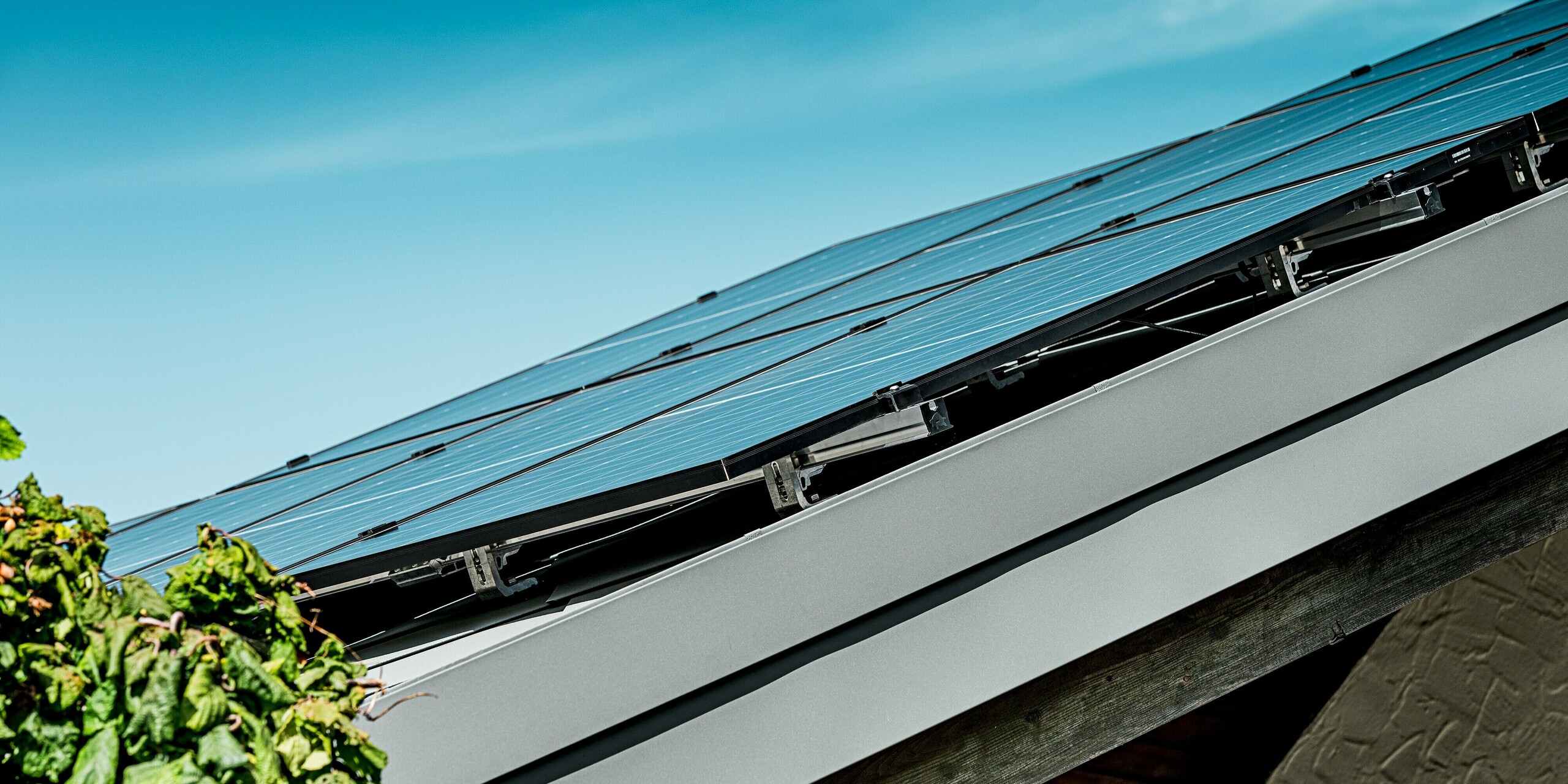 Das Bild zeigt eine seitliche Nahaufnahme der Photovoltaikanlage auf dem PREFA Aluminiumdach eines Einfamilienhauses. Die PV-Module sitzen präzise auf dem robusten PREFA Solarmontagesystem, das eine sichere und langlebige Befestigung gewährleistet. Die glatte Dachkante mit sauber ausgeführter Aluminium-Abschlussleiste bildet einen modernen Kontrast zum klaren Blau des Himmels. Im Vordergrund rankt grünes Laub, das dem technisch hochwertigen Aufbau eine natürliche Umgebung verleiht. Die Kombination aus stabilem PREFA Dach und effizienter Solartechnik wirkt besonders zukunftsorientiert.