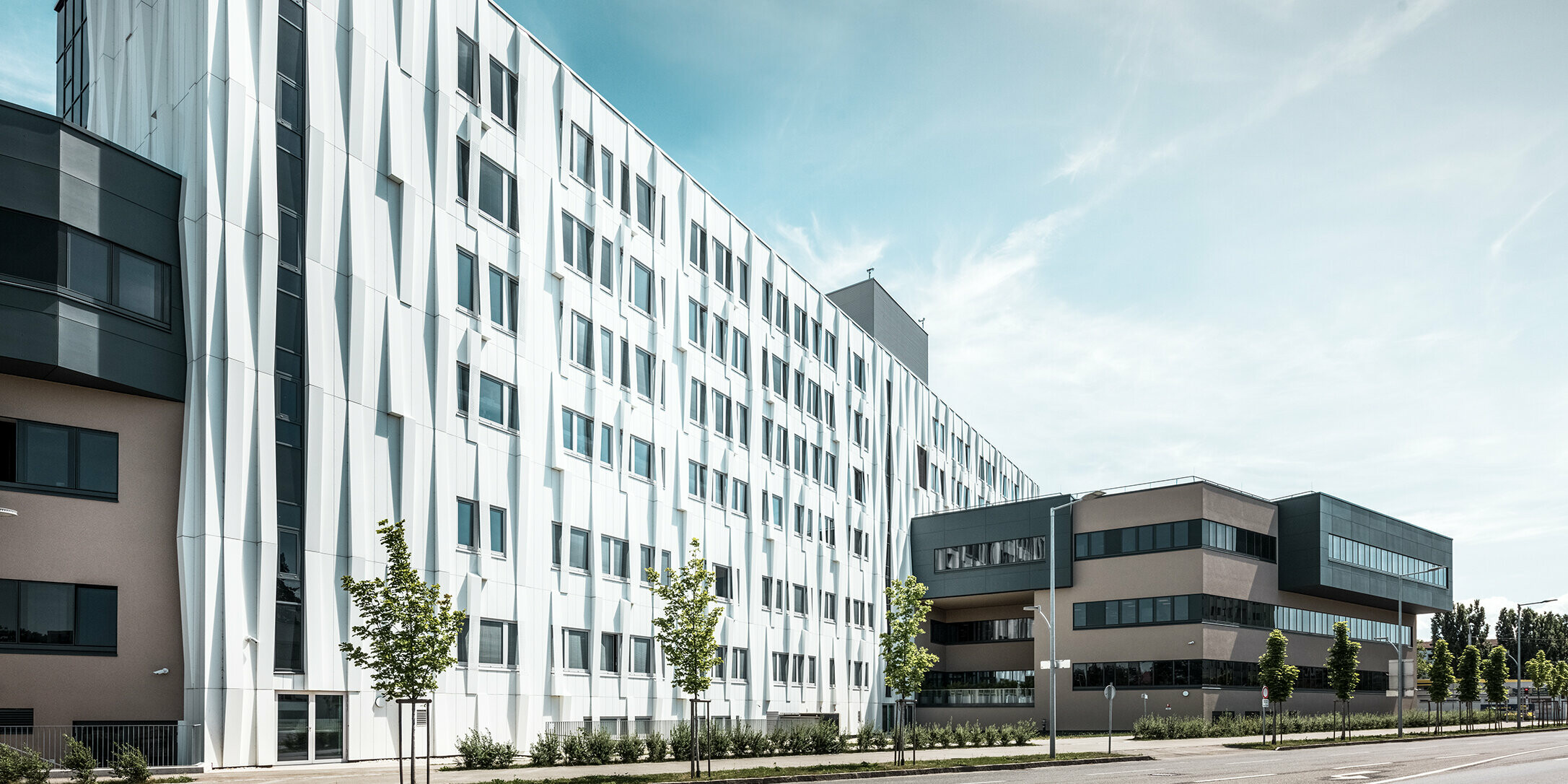 Seitliche Ansicht des Universitätsklinikums in St. Pölten, Niederösterreich. Die robuste Aluminiumfassade macht das Gebäude zu einem Blickfang in der Stadt. Im Vordergrund ist eine Straße mit einigen Bäumen zu sehen.