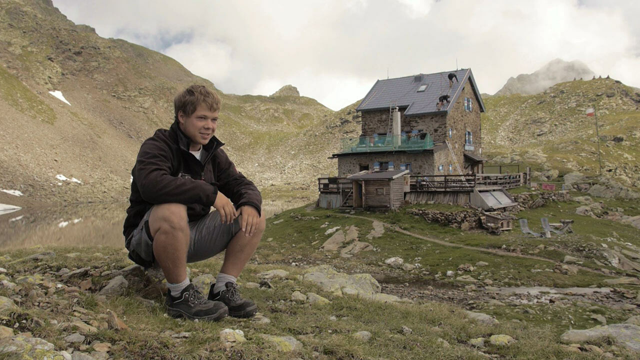 L’apprendista Jakob Schöttl è seduto su un masso. Sullo sfondo si può vedere il “Rifugio Forcella di Vallaga”, che è stata nuovamente rivestita con le tegole PREFA.