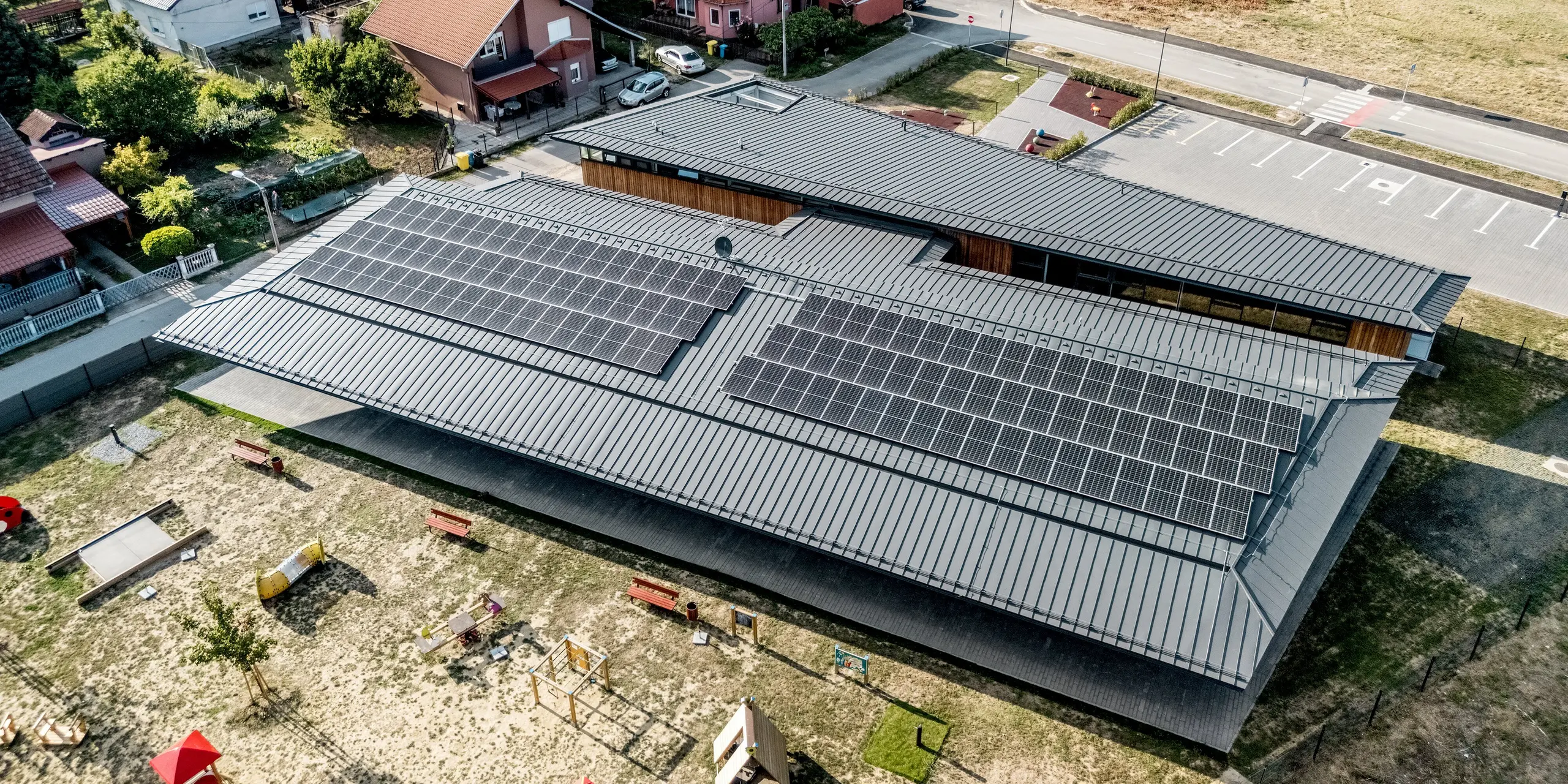 Drohnenaufnahme des Kindergartens Svarozic mit weitläufigem PREFALZ Aluminiumdach in P.10 Anthrazit und großflächiger Photovoltaikanlage. Die markante Dachstruktur überspannt das Gebäude mit Holzfassade und fügt sich harmonisch in die grüne Umgebung mit Spielplatz ein. PREFA Aluminium bietet langlebigen Witterungsschutz, geringes Gewicht und eine moderne, zeitlose Optik.