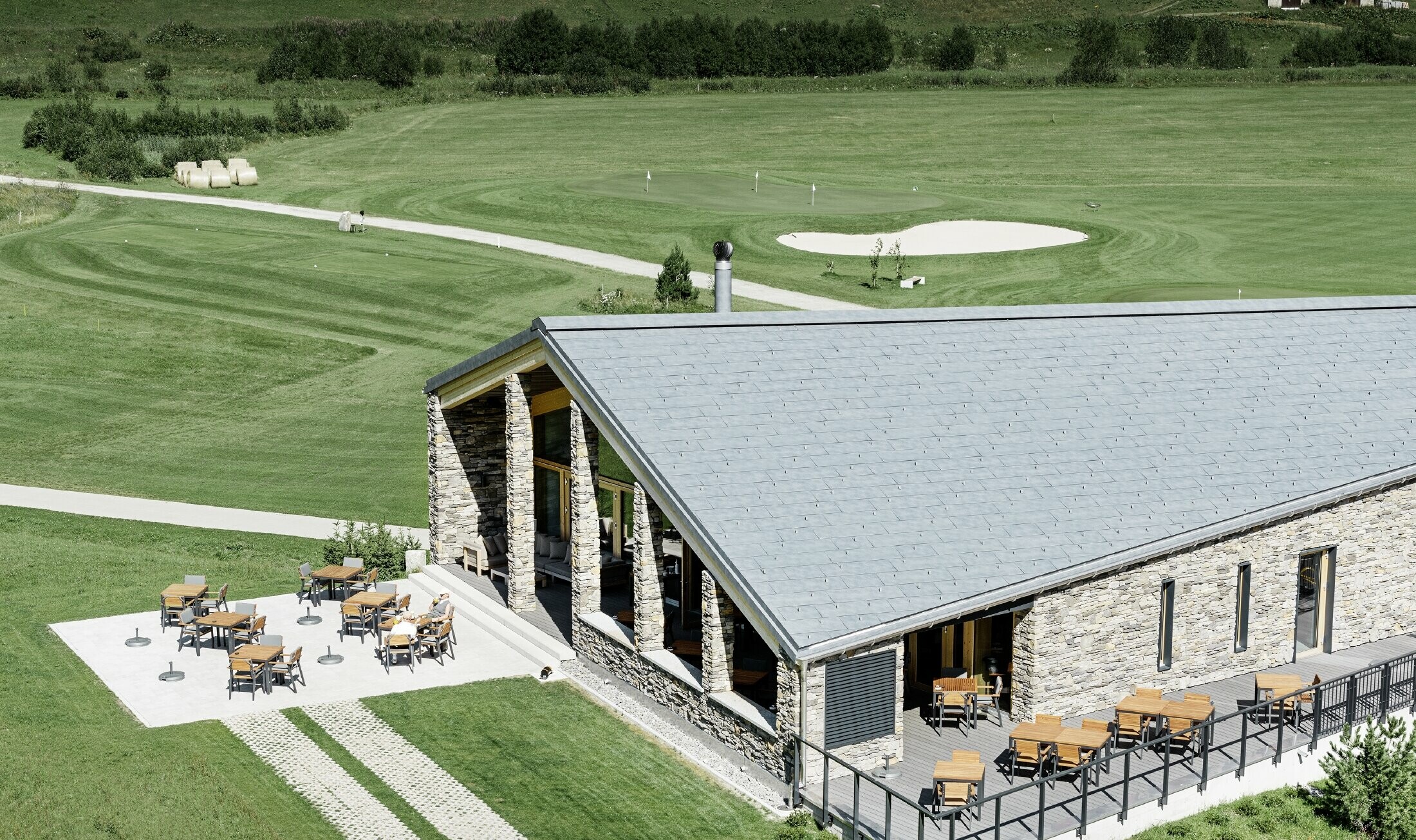 Il moderno punto di ritrovo del campo da golf a Andermatt con una facciata in pietra e il pannello per coperture FX.12 PREFA in alluminio in grigio pietra