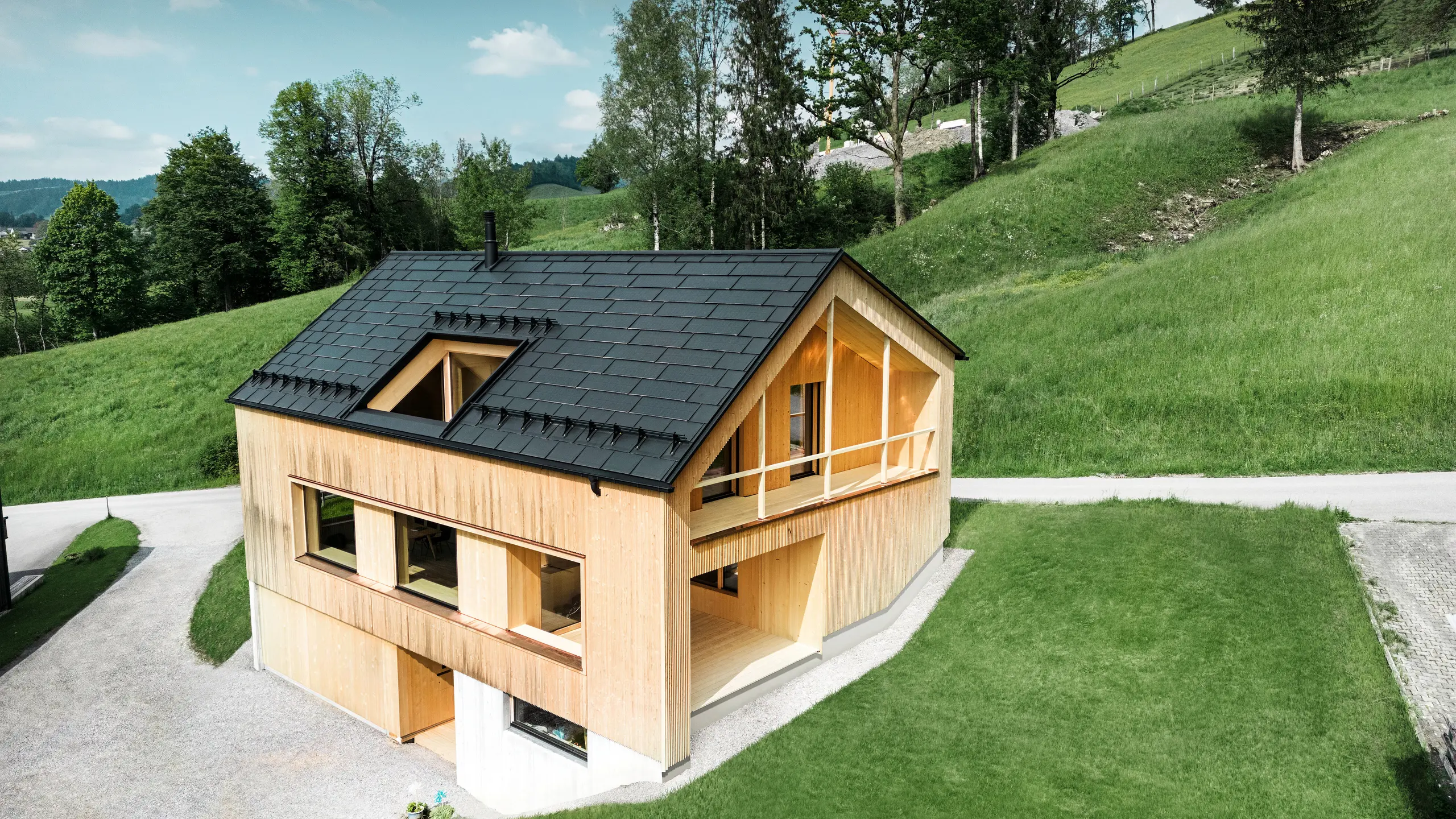 L'immagine mostra una pittoresca casa ecologica a Egg, immersa nel verde delle colline, con un tetto PREFA Solar sostenibile. Il sistema di copertura R.16 nero di alta qualità PREFA e le tegole fotovoltaiche PREFA si inseriscono armoniosamente nel paesaggio rurale e sottolineano la bellezza naturale dell'ambiente circostante. Il Sistema PREFA Solar per tetti non è solo un impegno verso le energie rinnovabili, ma anche un elemento di design che completa con stile la moderna facciata in legno e le linee pulite dell'edificio. Questa casa è un modello di edilizia ecologica e un'ispirazione per un'architettura orientata al futuro.
