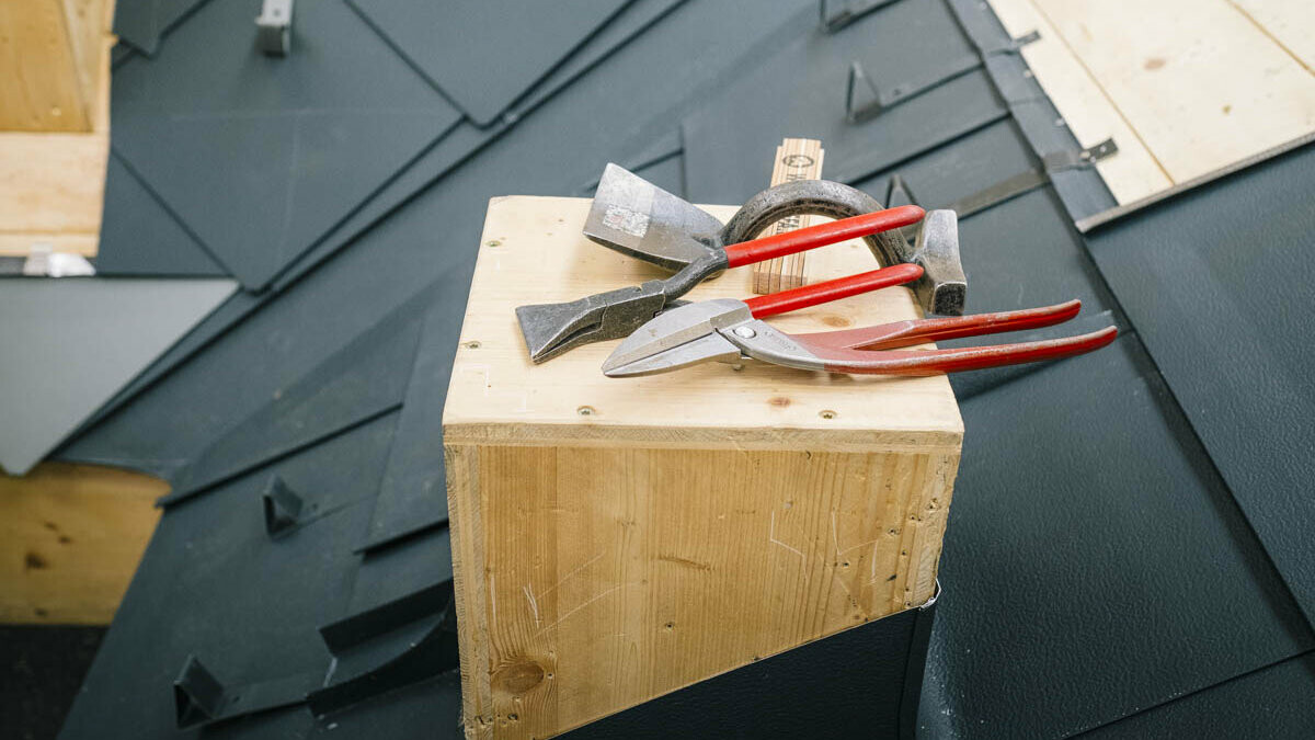 Dettaglio dalla PREFA Academy Italia: su un elemento di esercitazione in legno è disposta una selezione di utensili manuali, tra cui cesoie per lamiera, pinza per aggraffatura, martello e strumento di tracciatura, tipici utensili per la lavorazione dei sistemi per copertura PREFA. Sullo sfondo è visibile un'area di formazione coperta con tegole PREFA.