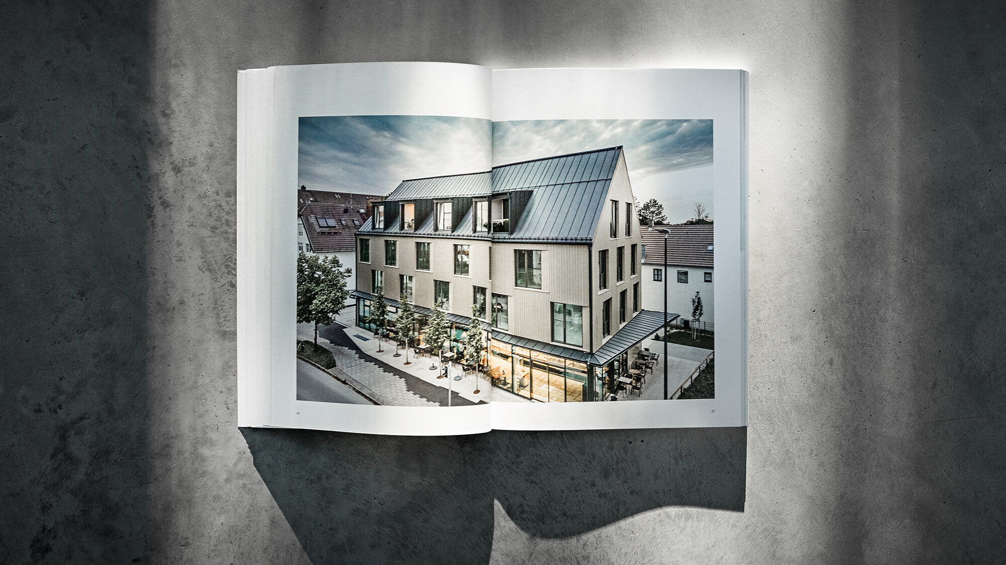 Libro PREFARENZEN 2024 aperto, su sfondo grigio, con due pagine su cui è raffigurato l'edificio commerciale e residenziale di Feldkirchen progettato dallo studio Heigl.