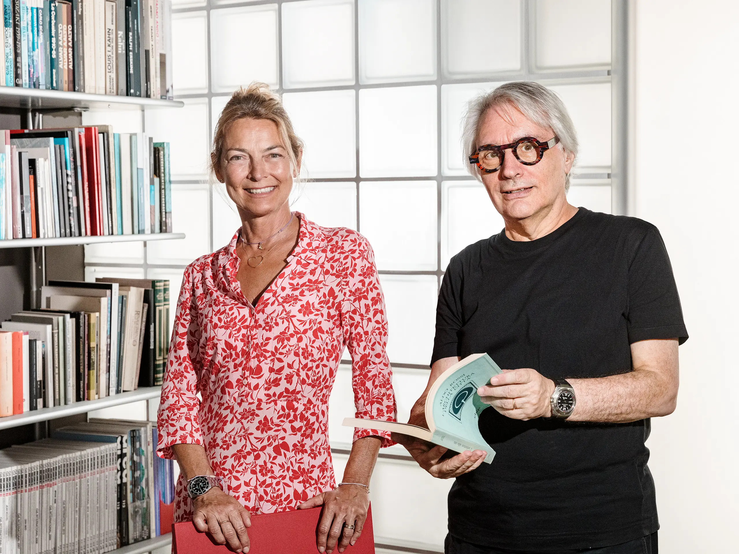 L'architetto bolzanino Claudio Lucchin e la sua socia Daniela Varnier posano davanti a una moderna parete in vetrocemento che rifrange la luce in modo suggestivo. Claudio Lucchin è vestito completamente di nero e tiene in mano un libro. Daniela Varnier indossa un top rosso acceso e sorride amichevolmente. Questo scenario elegante simboleggia innovazione e trasparenza. La scena rappresenta l'architettura orientata al futuro, come quella realizzata nella caserma “Tempesti” a Corvara in Alto Adige.