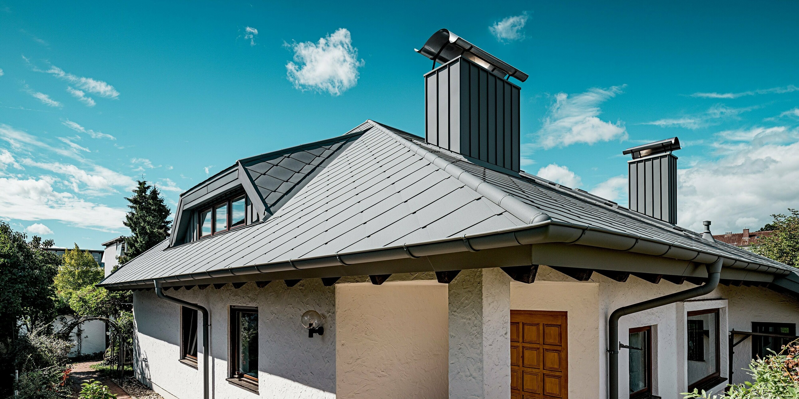 Das Bild zeigt den Eingangsbereich eines Einfamilienhauses in Ingolstadt mit einem markanten Walmdach aus PREFA Dachrauten 44 × 44 in P.10 Hellgrau. Die präzise verlegte Rautenstruktur und die saubere Linienführung betonen die hochwertige Aluminiumoptik. Die elegant verkleideten Kaminzüge und die farblich abgestimmte Dachentwässerung setzen weitere Akzente. Das robuste PREFA Aluminium sorgt für extreme Langlebigkeit, Formstabilität und Witterungsbeständigkeit – sichtbar in der klaren, modernen Dachgestaltung unter einem strahlend blauen Himmel.