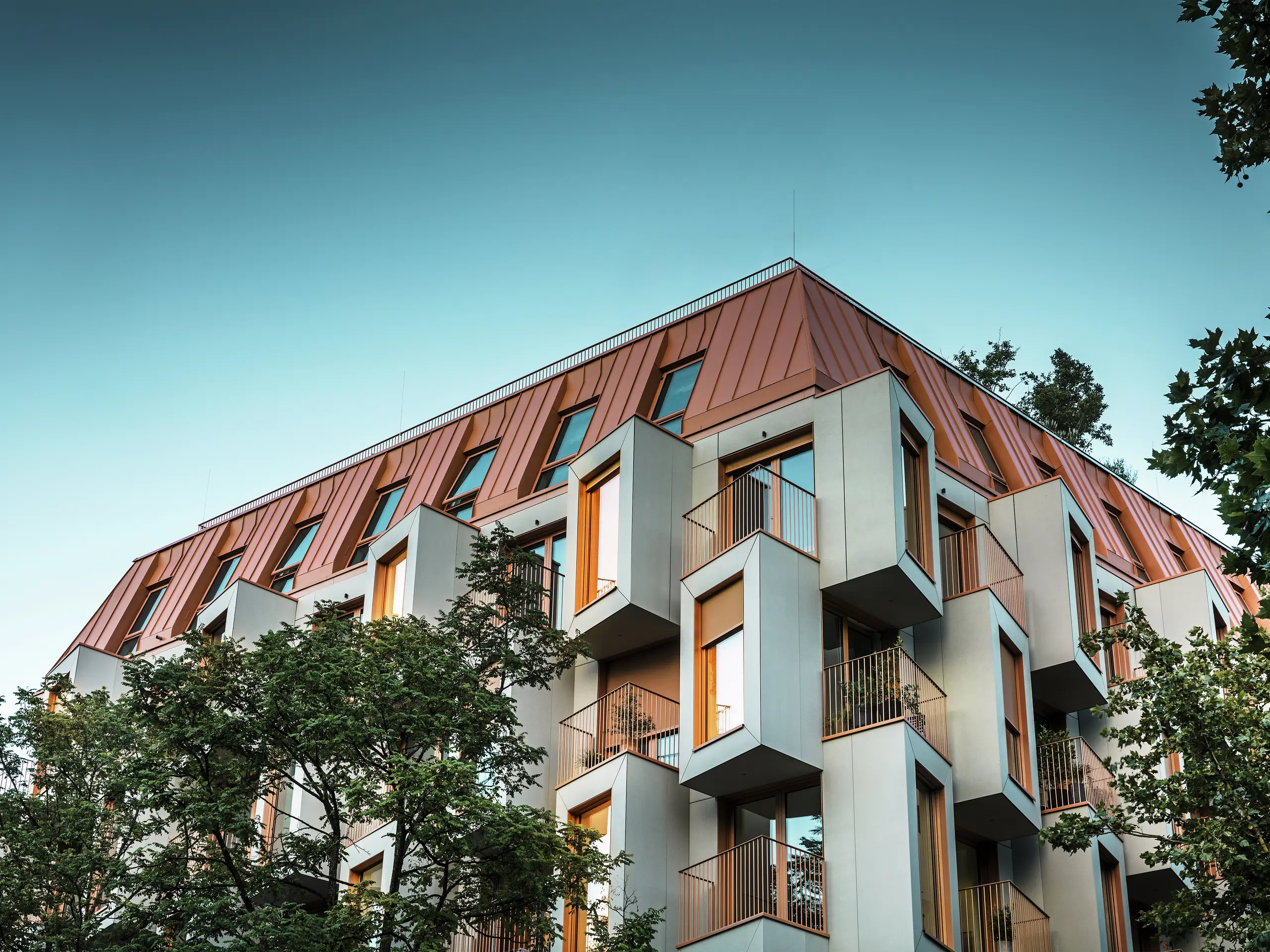 Facciata architettonicamente straordinaria del progetto residenziale Van B a Monaco di Baviera, con suggestive finestre a bovindo e tetto in alluminio PREFALZ nel colore speciale IGP Orange HDP.  Al crepuscolo, la vista dal basso mostra le luci delle finestre. La moderna soluzione per il tetto combina la massima resistenza agli agenti atmosferici con un aspetto elegante e crea accenti colorati nello spazio urbano. L'interazione tra il calcestruzzo in fibra di vetro, gli infissi in rame arancione e il tetto in alluminio crea un involucro edilizio dinamico, durevole e sostenibile, ideale per un'edilizia orientata al futuro.