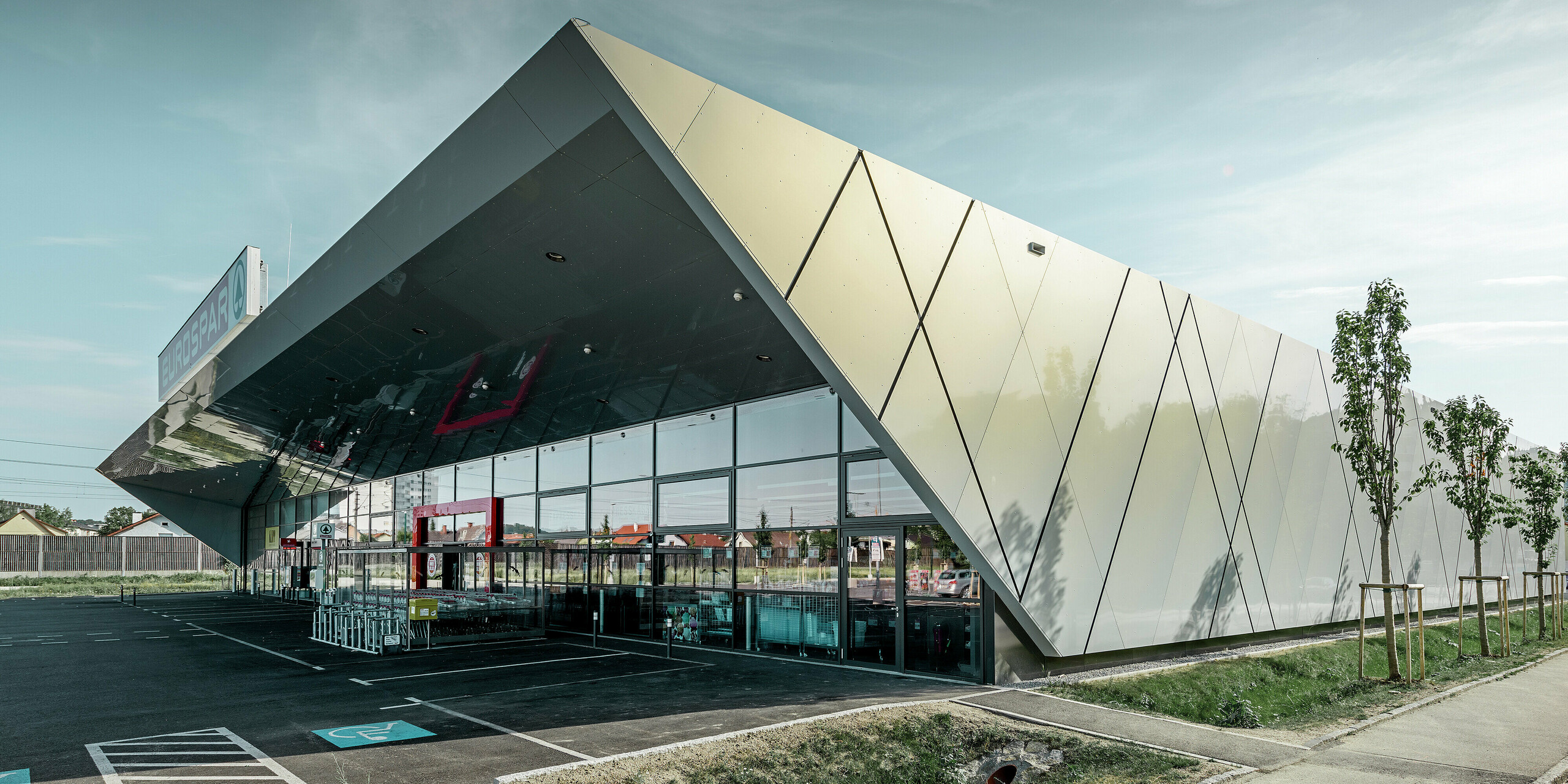 Zona d’ingresso della nuova filiale Eurospar di Wels con il moderno pannello per facciata PREFA in argento dorato; i pannelli compositi in alluminio brillano al sole in diversi colori a seconda della prospettiva.