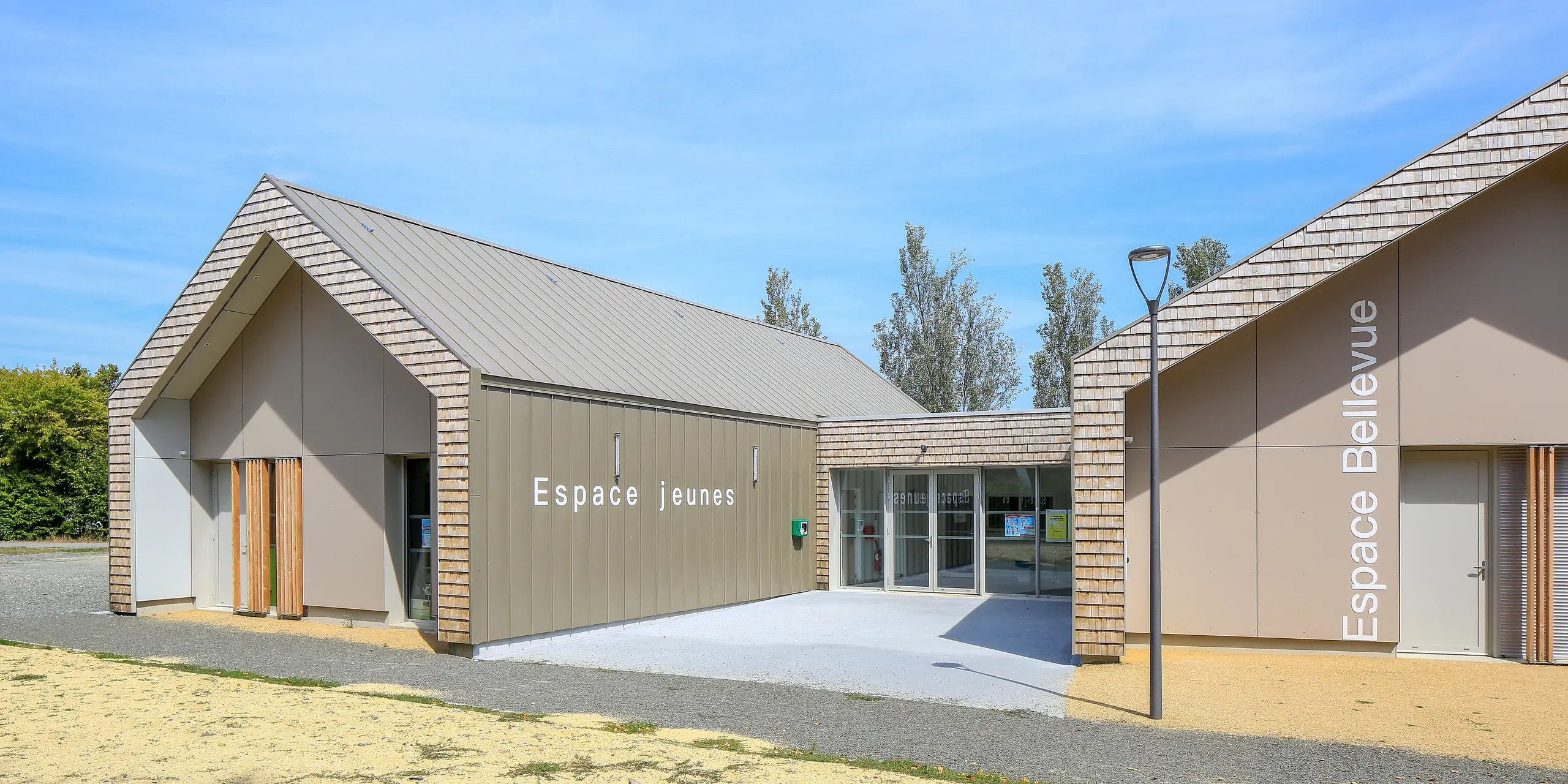Salles Espace Bellevue in Mûrs-Erigné, Frankreich, mit den Schriftzügen 'Espace Jeunes' und 'Espace Bellevue' vor dem Veranstaltungssaal. Das PREFALZ Dach- und Fassadensystem in Bronze schmückt große Teile des modernen Jugendzentrums. Die witterungsbeständige Aluminium-Stehfalzverkleidung von PREFA bietet eine langlebige und moderne Lösung für Bauvorhaben. Die Kombination aus Aluminiumverkleidung und natürlicher Holzoptik fügt sich harmonisch in die Umgebung ein.
