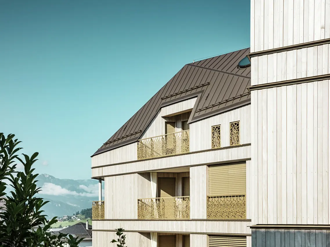 Primo piano di un moderno edificio residenziale a Liebwylen, in Svizzera, con tetto in alluminio PREFALZ di colore marrone.  La facciata in legno è completata da ringhiere dal design artistico. Sullo sfondo le montagne e il cielo azzurro, mentre in primo piano è visibile parte della vegetazione circostante.