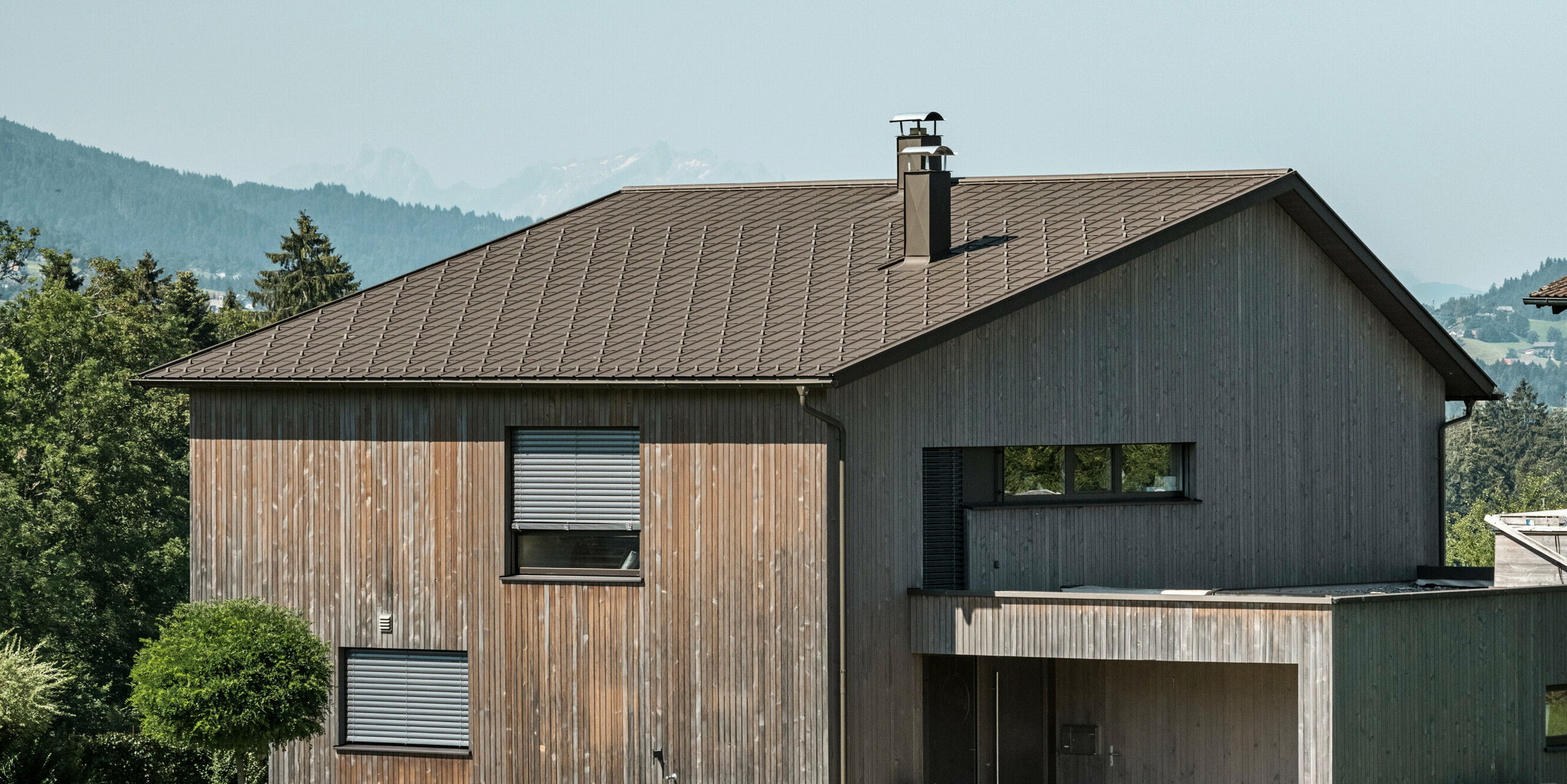 Vista laterale di una casa unifamiliare contemporanea a Krumbach con una combinazione di facciata in legno naturale e tetto in alluminio PREFA di lunga durata in P.10 marrone. L'armoniosa combinazione di colori mostra l'elegante integrazione di design moderno e materiali tradizionali, mentre le tegole PREFA assicurano un design del tetto estetico e funzionale.