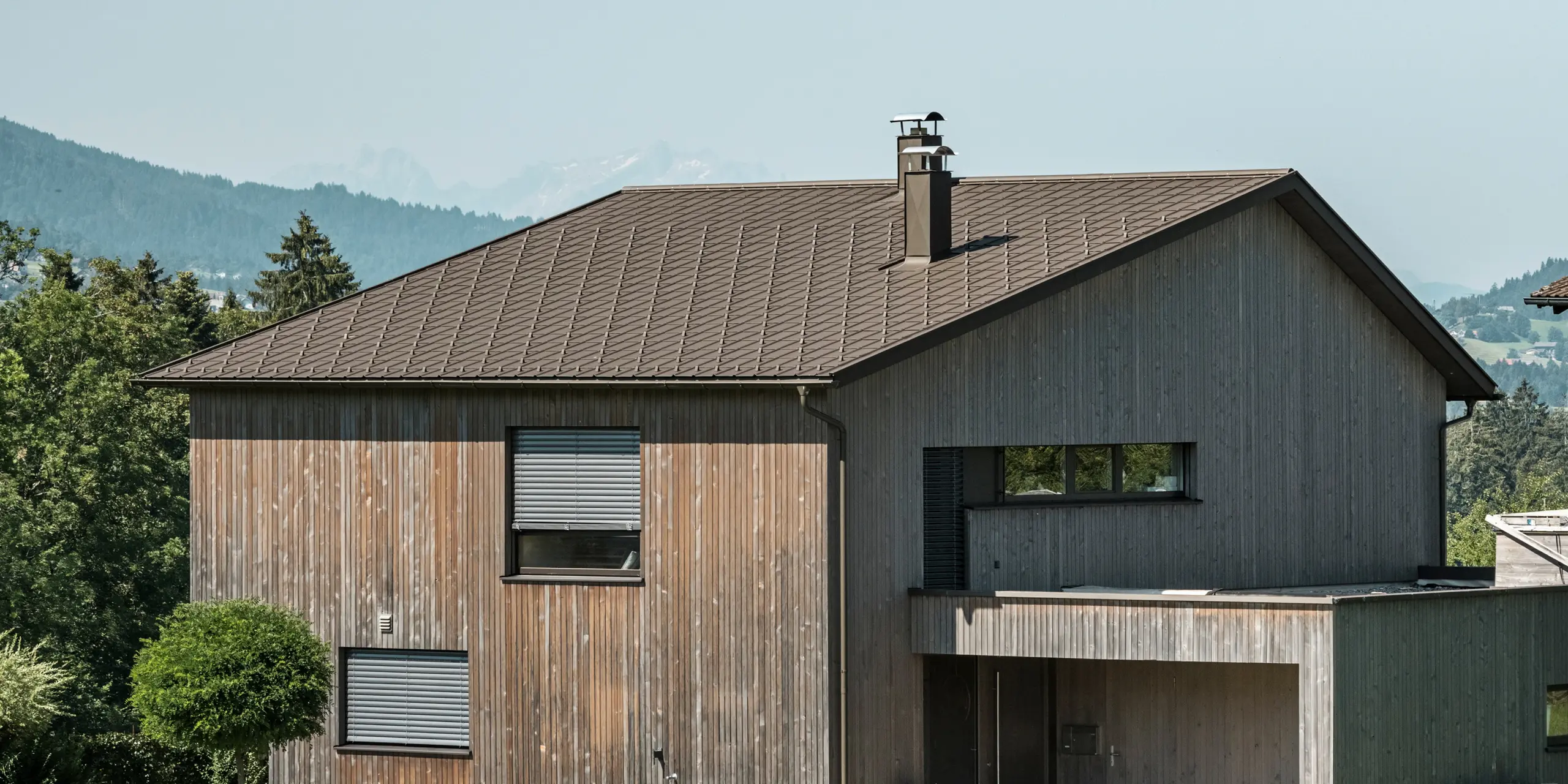 Seitenansicht eines zeitgenössischen Einfamilienhauses in Krumbach mit einer Kombination aus einer natürlichen Holzfassade und einem langlebigen PREFA Aluminiumdach in P.10 Braun. Die harmonische Farbgebung zeigt die elegante Integration von modernem Design und traditionellen Materialien, wobei die PREFA Dachplatten für eine ästhetische und funktionale Dachgestaltung sorgen.