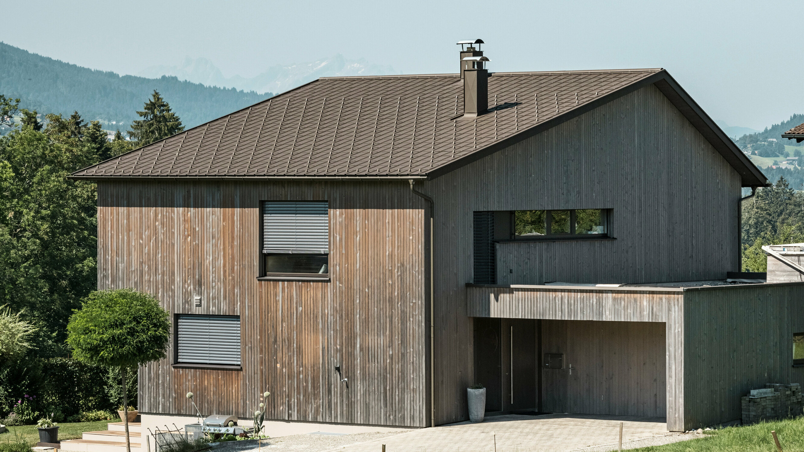 Vista laterale di una casa unifamiliare contemporanea a Krumbach con una combinazione di facciata in legno naturale e tetto in alluminio PREFA di lunga durata in P.10 marrone. L'armoniosa combinazione di colori mostra l'elegante integrazione di design moderno e materiali tradizionali, mentre le tegole PREFA assicurano un design del tetto estetico e funzionale.