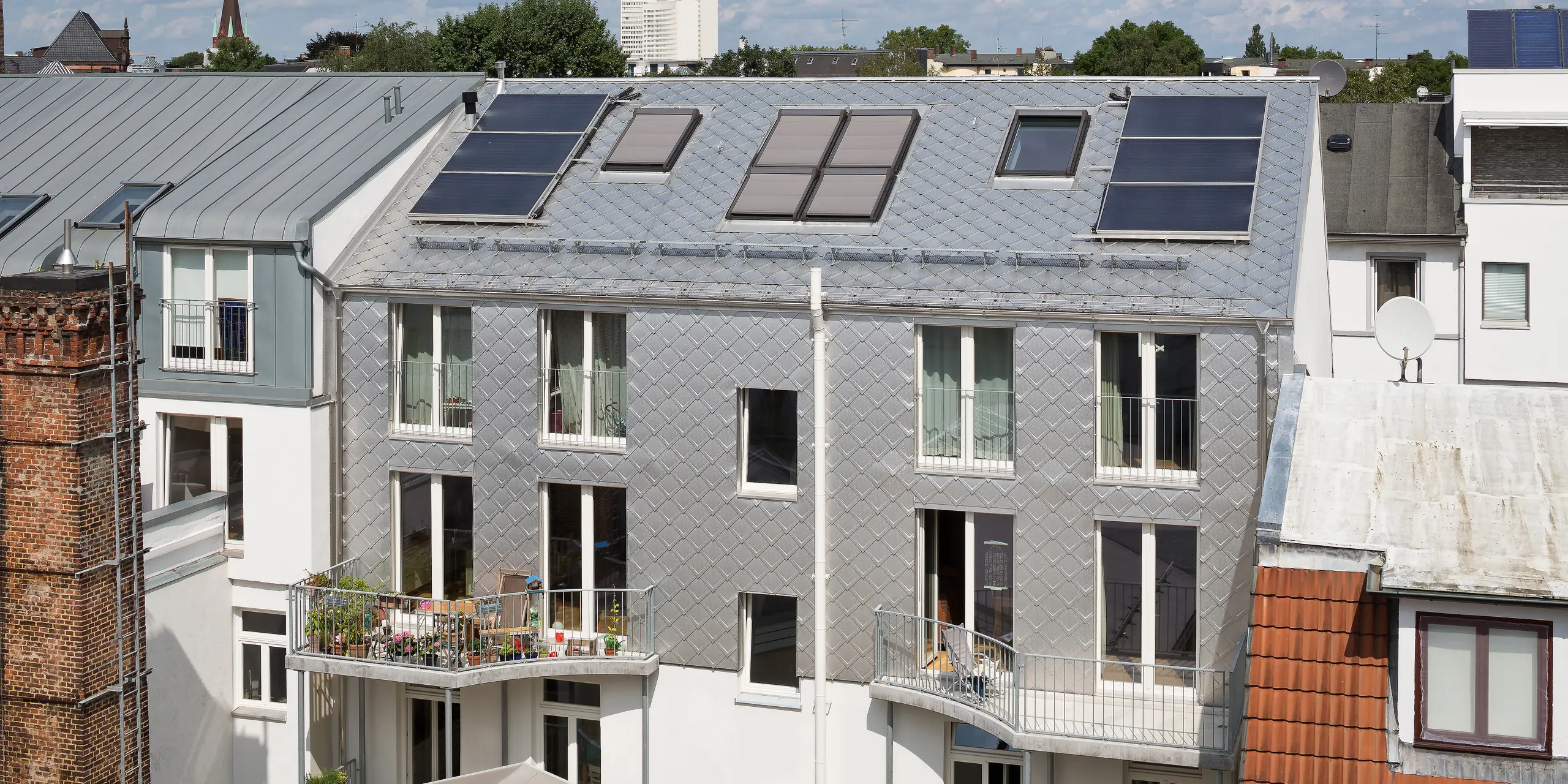 Das Dach eines eleganten Wohngebäudes in Hamburg besticht durch die Verwendung der PREFA Dachraute 29 × 29 in Naturblank. Die Rautenstruktur hebt sich beeindruckend von der Umgebung ab. Solarzellen auf dem Dach ergänzen das nachhaltige Design, während die Balkone der unteren Etagen das Gebäude noch einladender wirken lassen.