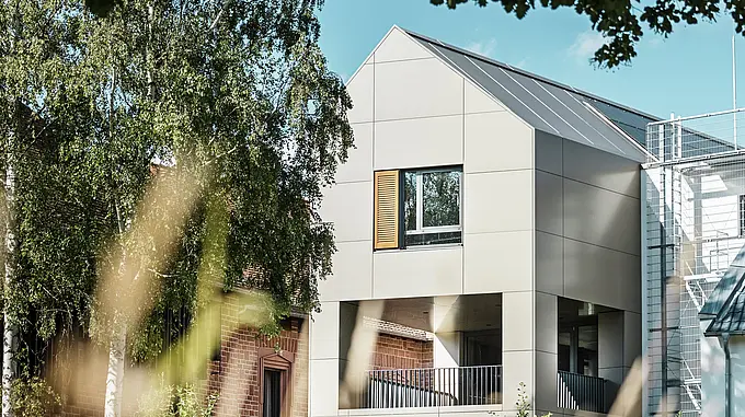 L'immagine mostra il moderno municipio di Alzenau, la cui facciata è realizzata con pannelli compositi in alluminio PREFA in un'elegante tonalità bronzo. Il tetto a punta e le grandi finestre sottolineano il carattere contemporaneo dell'edificio. In primo piano, alberi e aree verdi incorniciano l'edificio, mentre l'illuminazione soffusa e il cielo limpido sottolineano il moderno spazio urbano.