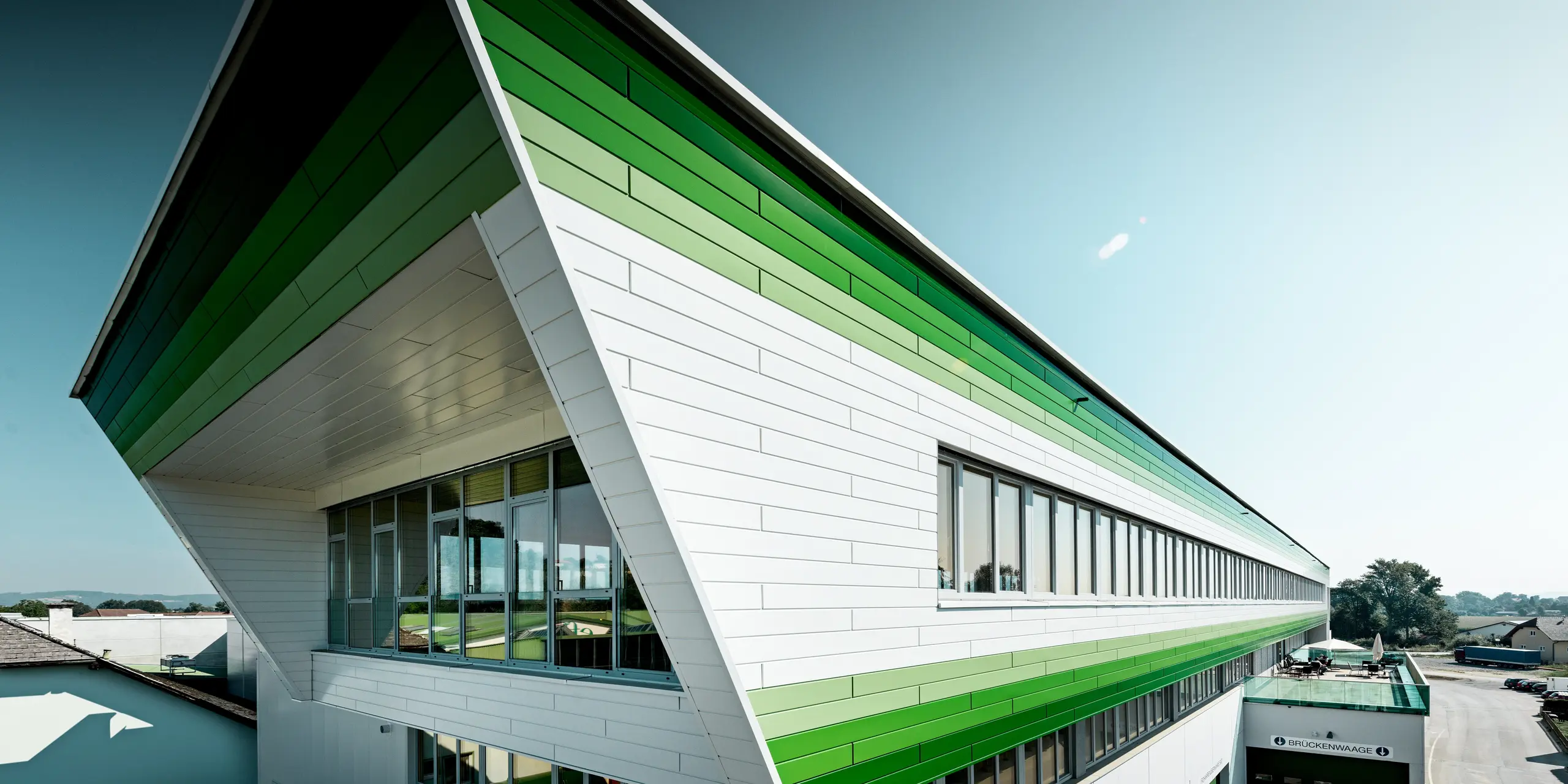 Weitläufige Ansicht des Firmengebäudes der efko Frischfrucht und Delikatessen GmbH mit markanter PREFA Siding-Fassade in P.10 Prefaweiß und individuellen grünen RAL-Sonderbeschichtungen. Die horizontalen Aluminiumprofile betonen die außergewöhnliche Gebäudelänge und schaffen ein dynamisches Fassadenbild. PREFA Aluminium steht für langlebige Qualität, hohe Witterungsbeständigkeit, Farbstabilität und eine wartungsarme Lösung für moderne Unternehmensarchitektur.