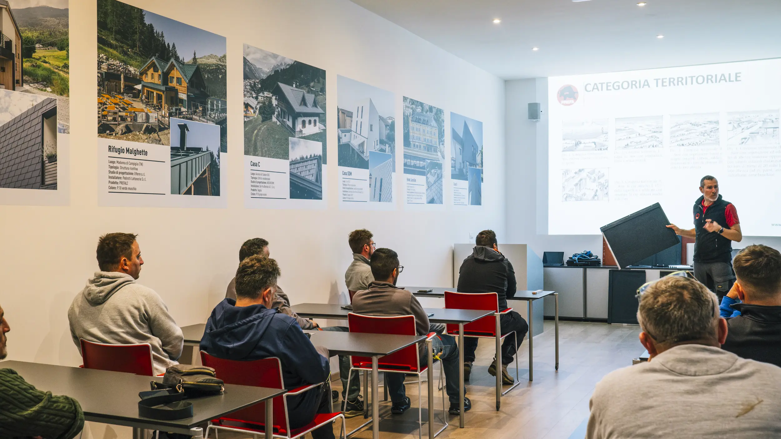 Ein Schulungsraum der PREFA Academy in Italien mit mehreren Teilnehmern, die an Tischen sitzen und einem Vortrag folgen. Ein Trainer steht vorne und hält eine PREFA-Dachplatte in der Hand.
