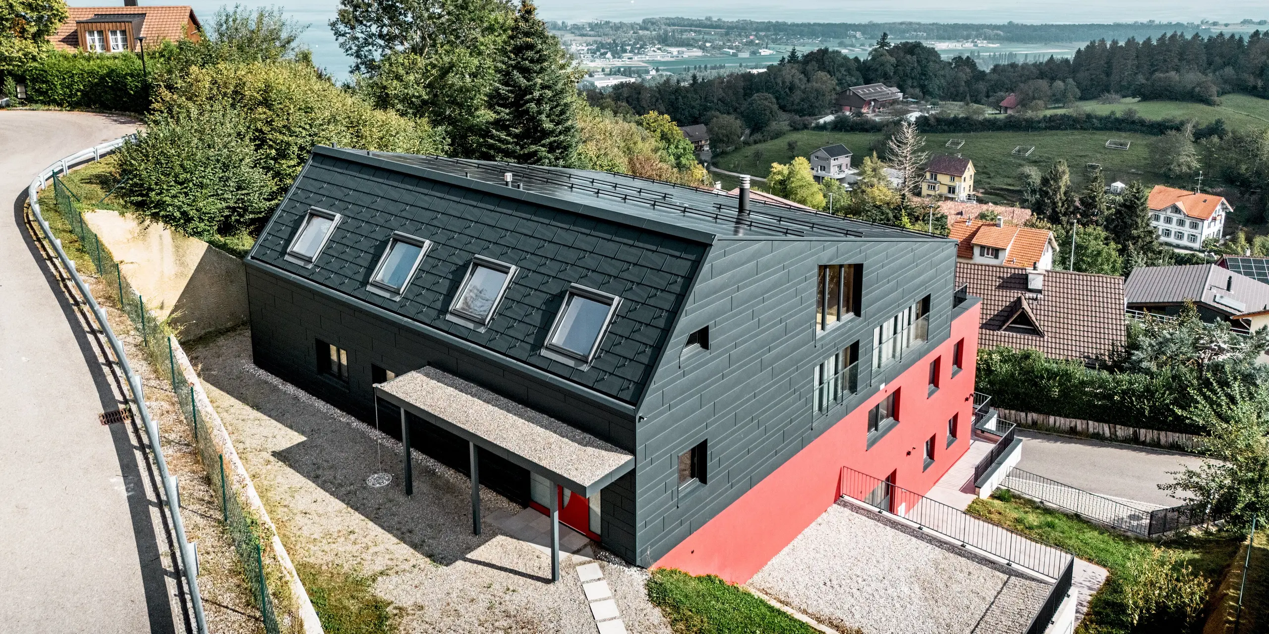 Hoch über dem Bodensee präsentiert sich das Weinwerk Wienacht-Tobel als architektonischer Blickfang. Die dunkle Gebäudehülle aus PREFA Dachpaneel FX.12 und Siding.X in P.10 Anthrazit kontrastiert eindrucksvoll mit dem roten Baukörper und der grünen Landschaft. Die langlebigen Aluminiumprodukte überzeugen durch Witterungsbeständigkeit, geringe Wartung und eine präzise, moderne Gestaltung.