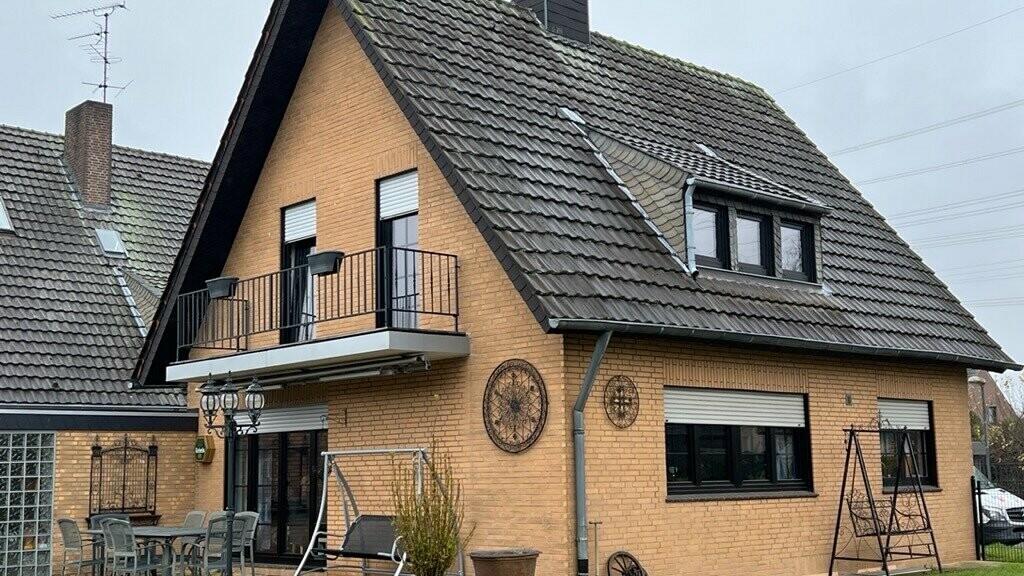 Einfamilienhaus aus gelbem Klinker mit steilem Satteldach und dunkler Dacheindeckung vor der Dachsanierung; auf der Gartenseite mit Balkon, Dachgaube, Terrasse mit Sitzmöbeln und Rasenfläche im Vordergrund.