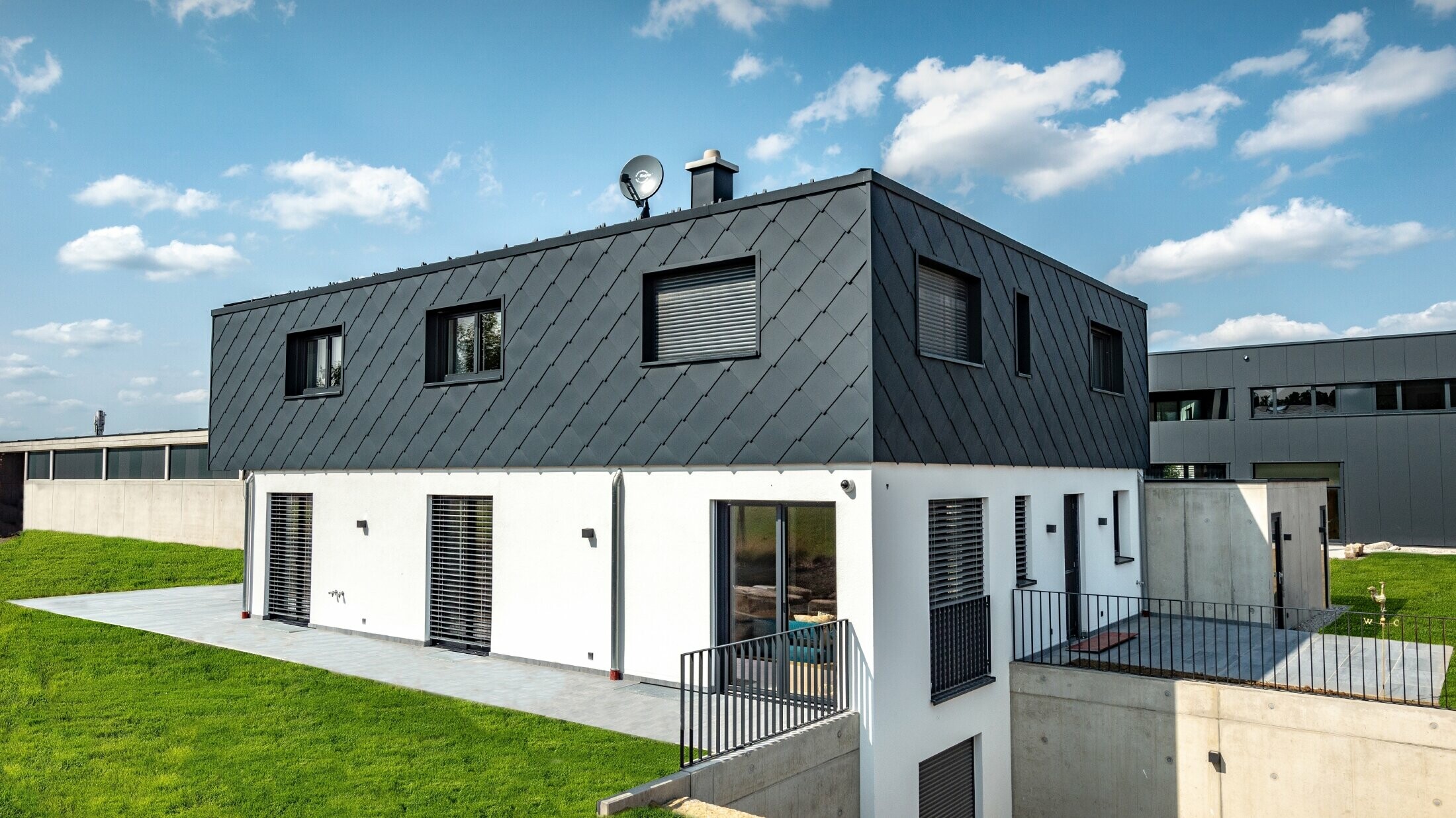 Semplice edificio di nuova costruzione con copertura piatta, il piano terra è intonacato di bianco, il piano superiore è rivestito con la scaglia PREFA in antracite.