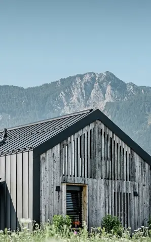 Moderner, funktionaler Wohnbau mit einer Kombination aus Holzfassade und PREFA Band- und Blechmaterial für das Dach. Die klare Architektur wird durch die elegante, metallische Dach- und Fassadenverkleidung unterstrichen, die ideal für anspruchsvolle Projekte ist. Das Bild veranschaulicht die vielseitigen Anwendungsmöglichkeiten der PREFA Produkte im Bereich Bänder und Bleche.