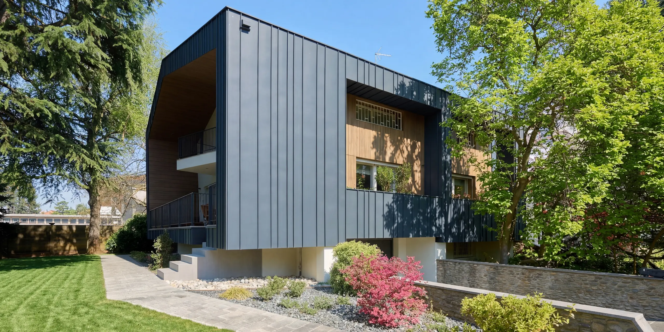 Ansicht der Villa Castellanza mit markanter PREFALZ Fassade in P.10 Anthrazit. Die klaren Stehfalzlinien umrahmen warme Holzelemente und verleihen dem Gebäude eine moderne, elegante Ausstrahlung. Umgeben von üppigem Grün zeigt die langlebige PREFA Aluminiumhülle ihre architektonische Stärke und verbindet Design, Witterungsbeständigkeit und nachhaltige Qualität harmonisch.