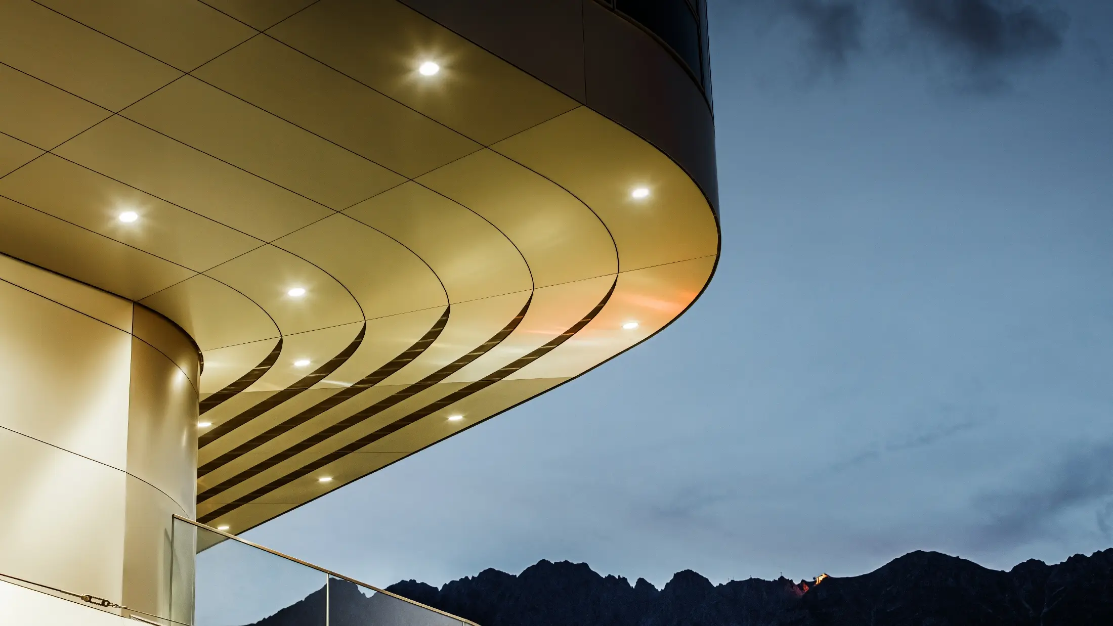 L'immagine mostra un impressionante dettaglio architettonico di un pannello composito PREFA PEMA in alluminio spazzolato. La lastra è leggermente bombata e presenta linee eleganti e fluide, che risaltano particolarmente la sera grazie a un'illuminazione mirata. La struttura chiara e l'aspetto raffinato dell'alluminio della lastra completano il design moderno della facciata e offrono una soluzione elegante e contemporanea per un'architettura di alta qualità.