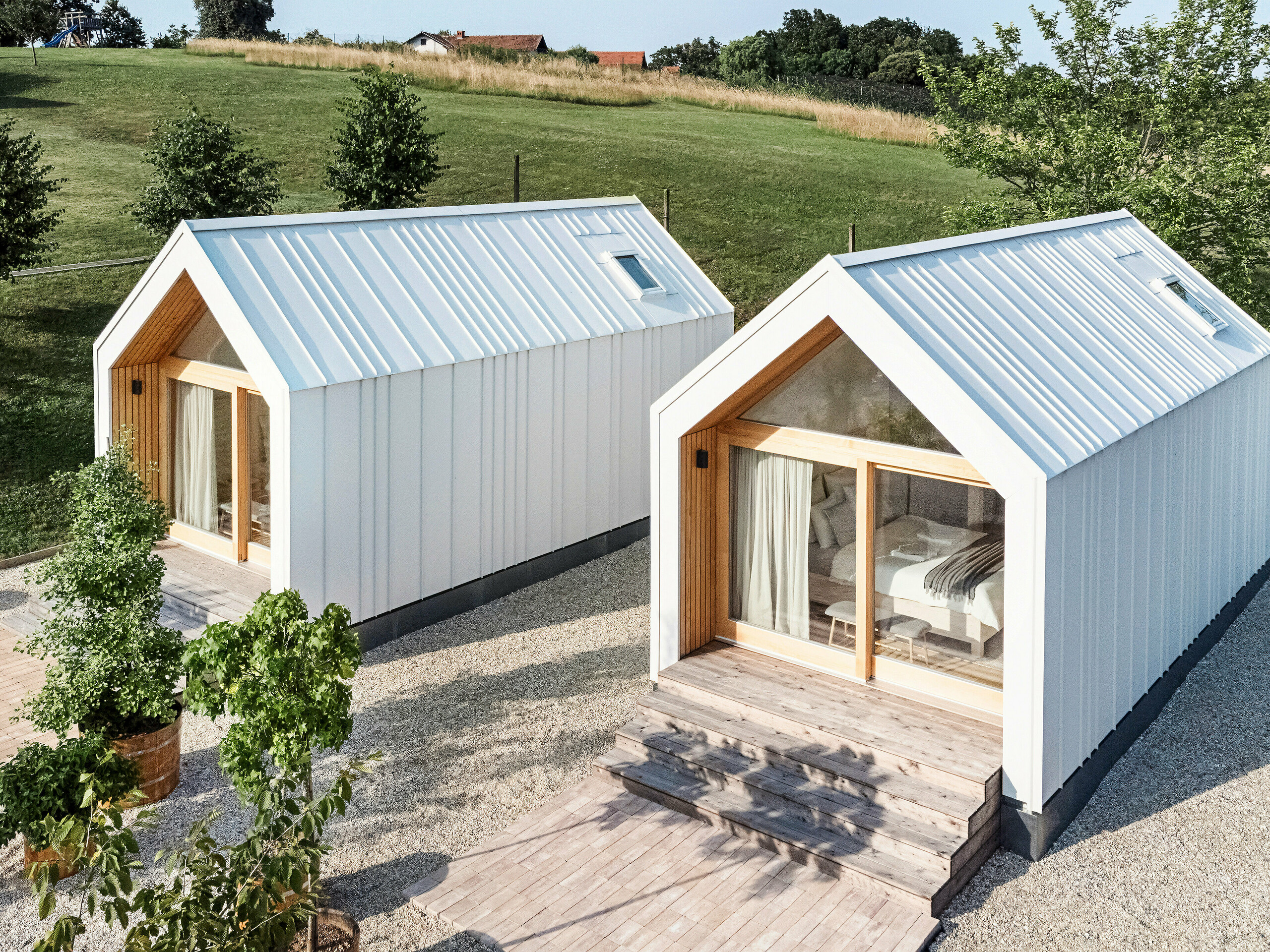 Blick auf die Vorderseite der beiden Tiny Häuser der Ferienanlage 'Pri Momi' in Kančevci, Slowenien. Die Mikrohäuser wurden in einer Holzrahmenbauweise erreichtet und mit einer robusten PREFALZ Dach- und Fassadenverkleidung in P.10 Reinweiß vollendet. Die modernen, minimalistischen Bauwerke kombinieren eine langlebige und zugleich ästhetische Aluminiumhülle mit großen Glasfronten und naturbelassenem Holz, eingebettet in eine grüne Landschaft. Die großzügigen Holzstufen führen zu den Eingängen, die eine gemütliche und einladende Atmosphäre schaffen.
