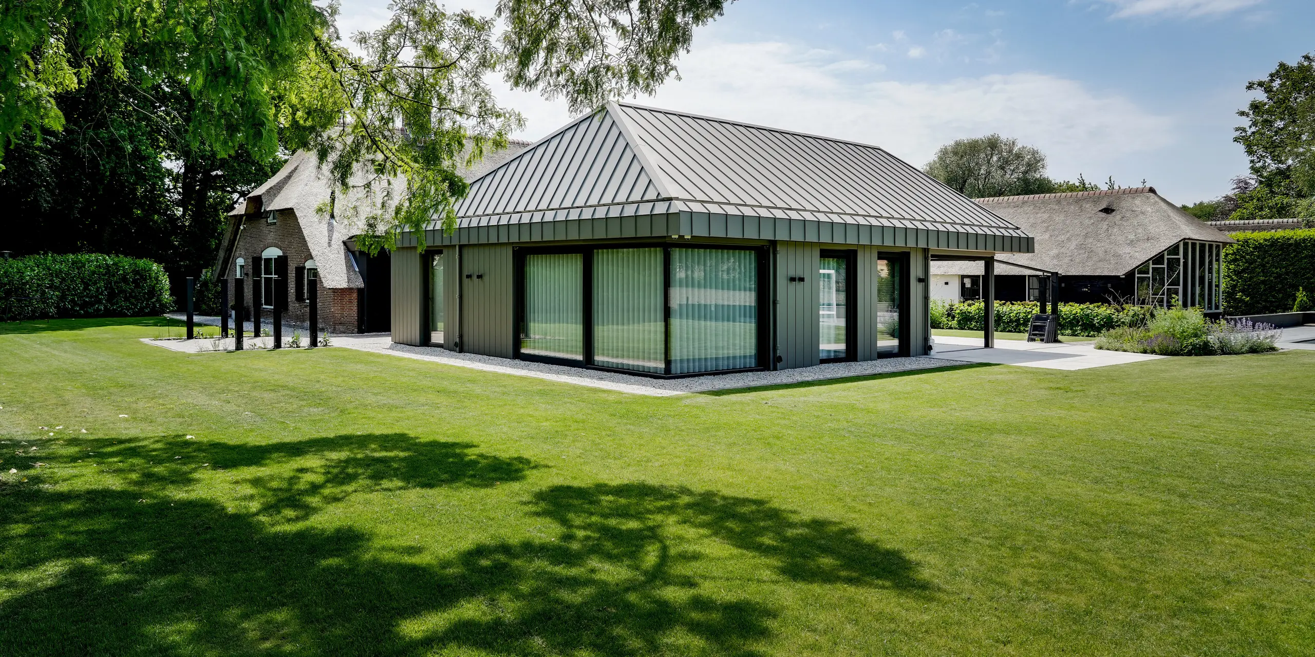 Eindrucksvolle Gesamtansicht des Einfamilienhauses in Putten mit modernem PREFALZ Anbau und historischem Reetdach. Die elegante Aluminiumhülle in PREFABRONZE verbindet Dach und Fassade zu einer klaren architektonischen Einheit. Großzügige Glasflächen, präzise Stehfalze und die weitläufige Gartenlandschaft unterstreichen die hochwertige Optik sowie die langlebigen, witterungsbeständigen Eigenschaften von PREFA Aluminium.