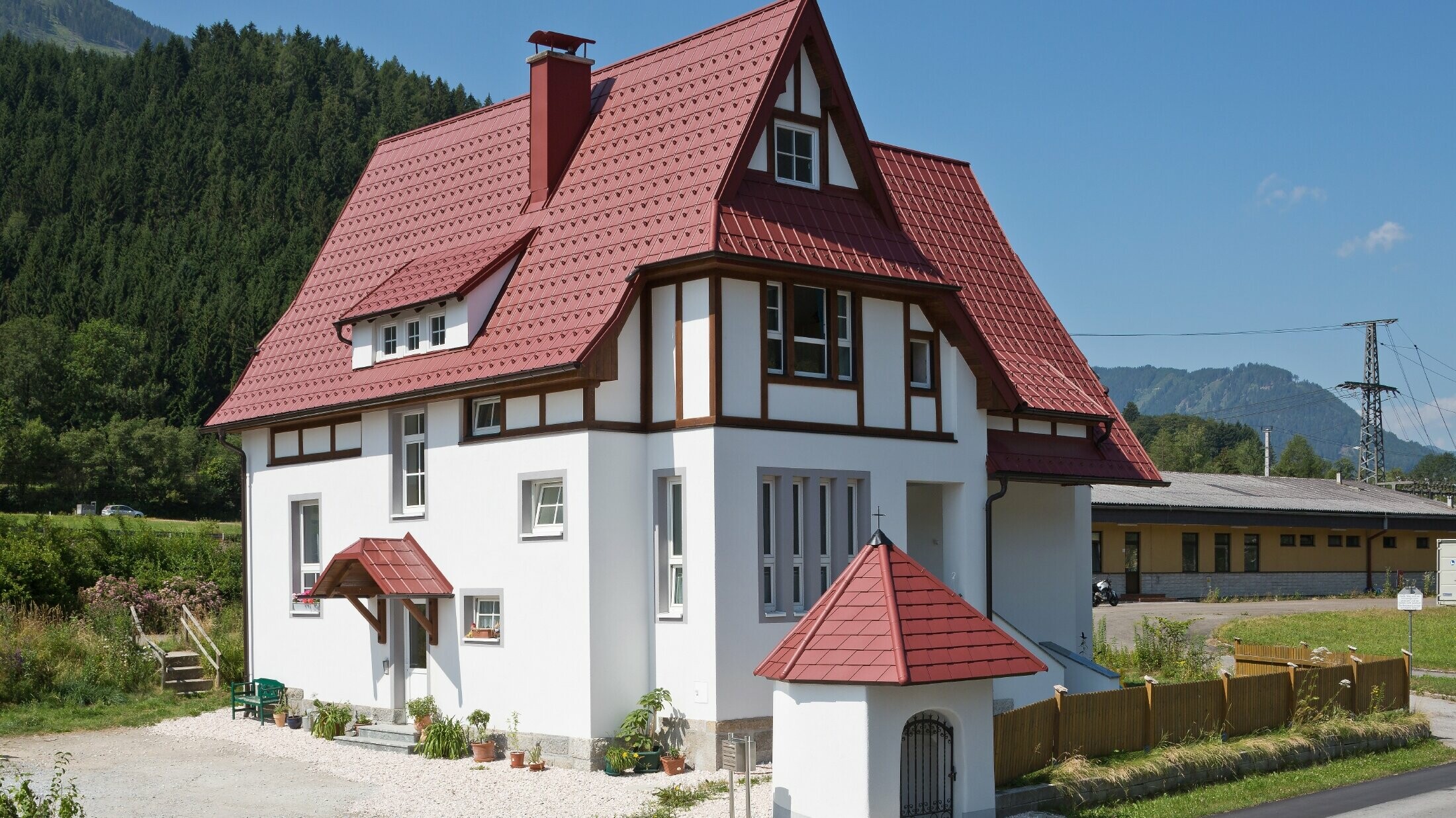 Verwinkeltes Steildach mit PREFA Dachplatte in P.10 Oxydrot mit vielen Details wie Gauben, Türmchen und Kamineinfassungen. Auch das Vordach und das Marterl vor dem Haus sind mit PREFA Blechdach eingedeckt.