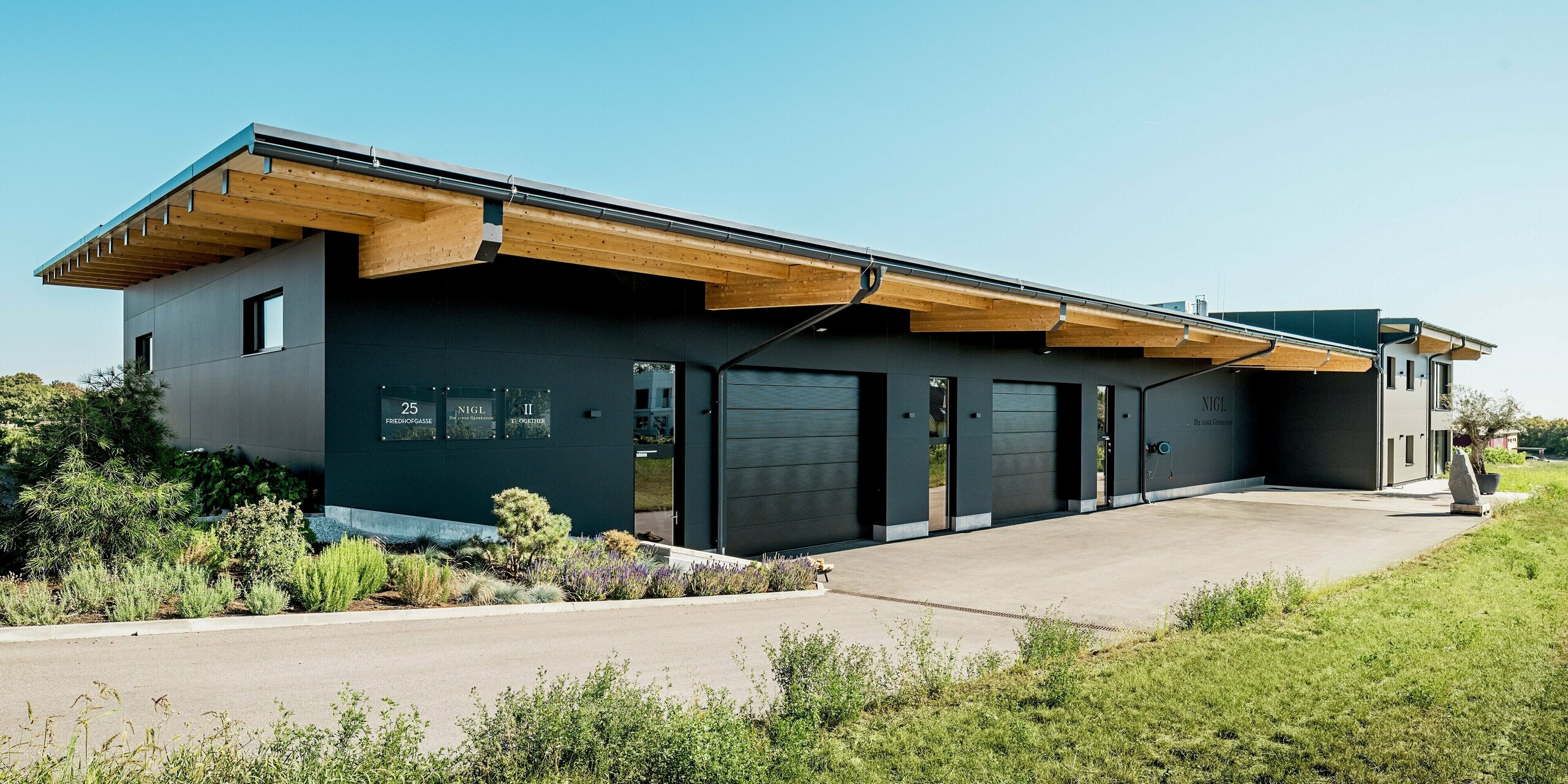 Modernes Betriebsgebäude des Weinguts Nigl in Perchtoldsdorf mit eindrucksvoller PREFA Aluminiumfassade in P.10 Schwarz. Die klar strukturierte Oberfläche der Aluminium Verbundplatten sorgt für ein elegantes, reduziertes Erscheinungsbild, das sich harmonisch in die Umgebung einfügt. Der Kontrast zwischen der dunklen Fassade, den natürlichen Holzelementen des weit auskragenden Flachldachs und dem saftig grünen Umfeld schafft eine markante architektonische Wirkung. PREFA Verbundplatten überzeugen hier nicht nur optisch, sondern auch durch hohe Witterungsbeständigkeit, Langlebigkeit und Wartungsfreiheit – ideal für moderne Gewerbebauten mit Stil und Funktion.