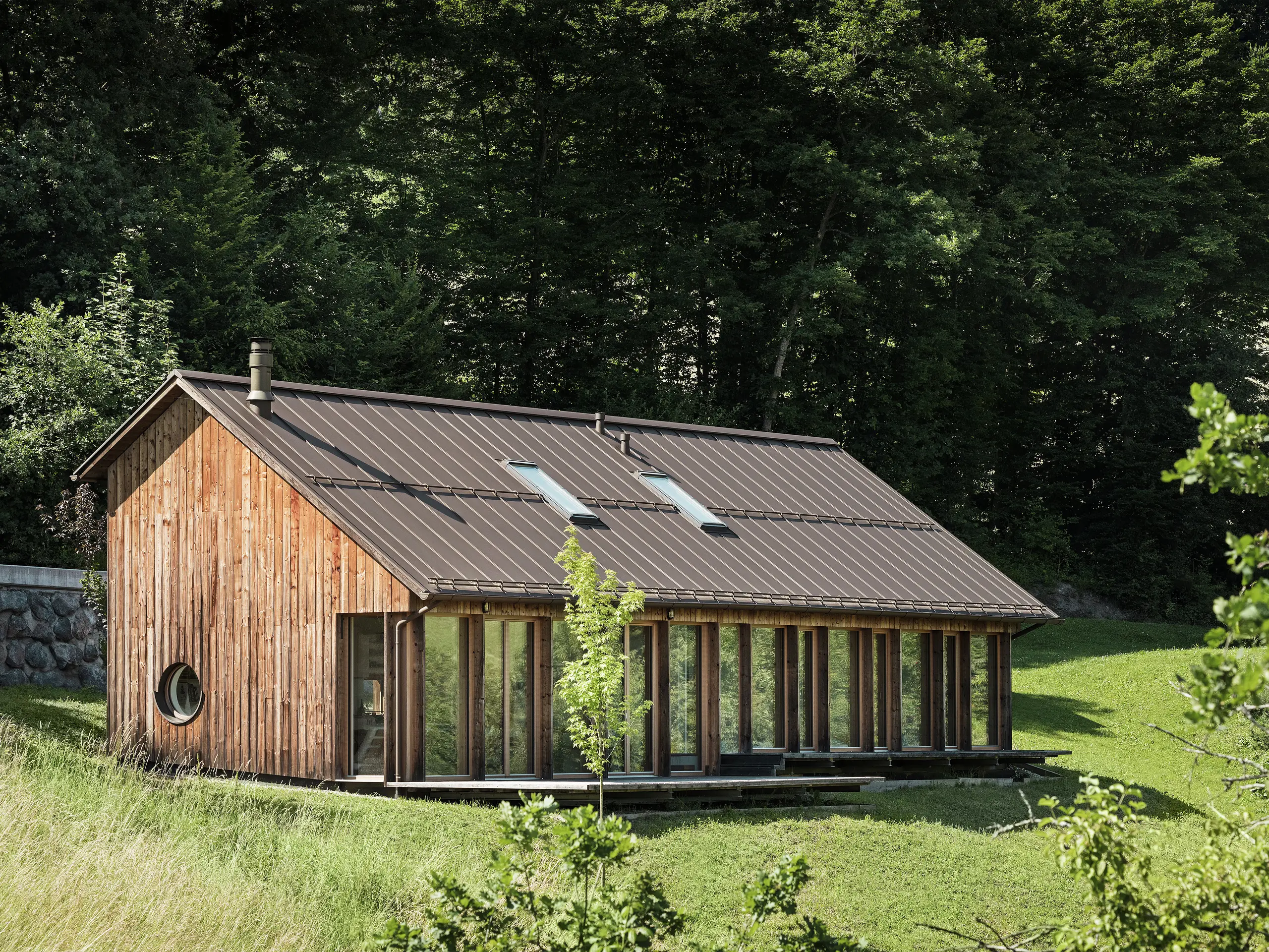 Ein Holzhaus mit großer Glasfassade und hochwertigem PREFALZ-Dach in der Farbe P.10 Braun – harmonisch eingebettet in die grüne Hügellandschaft von Vinje in Slowenien. Das robuste Aluminiumdach überzeugt durch Langlebigkeit, Korrosionsbeständigkeit und moderne Ästhetik. Integrierte Dachfenster sorgen für natürliches Licht und Energieeffizienz im Wohnraum. Das Haus ist von zahlreichen Bäumen umgeben.