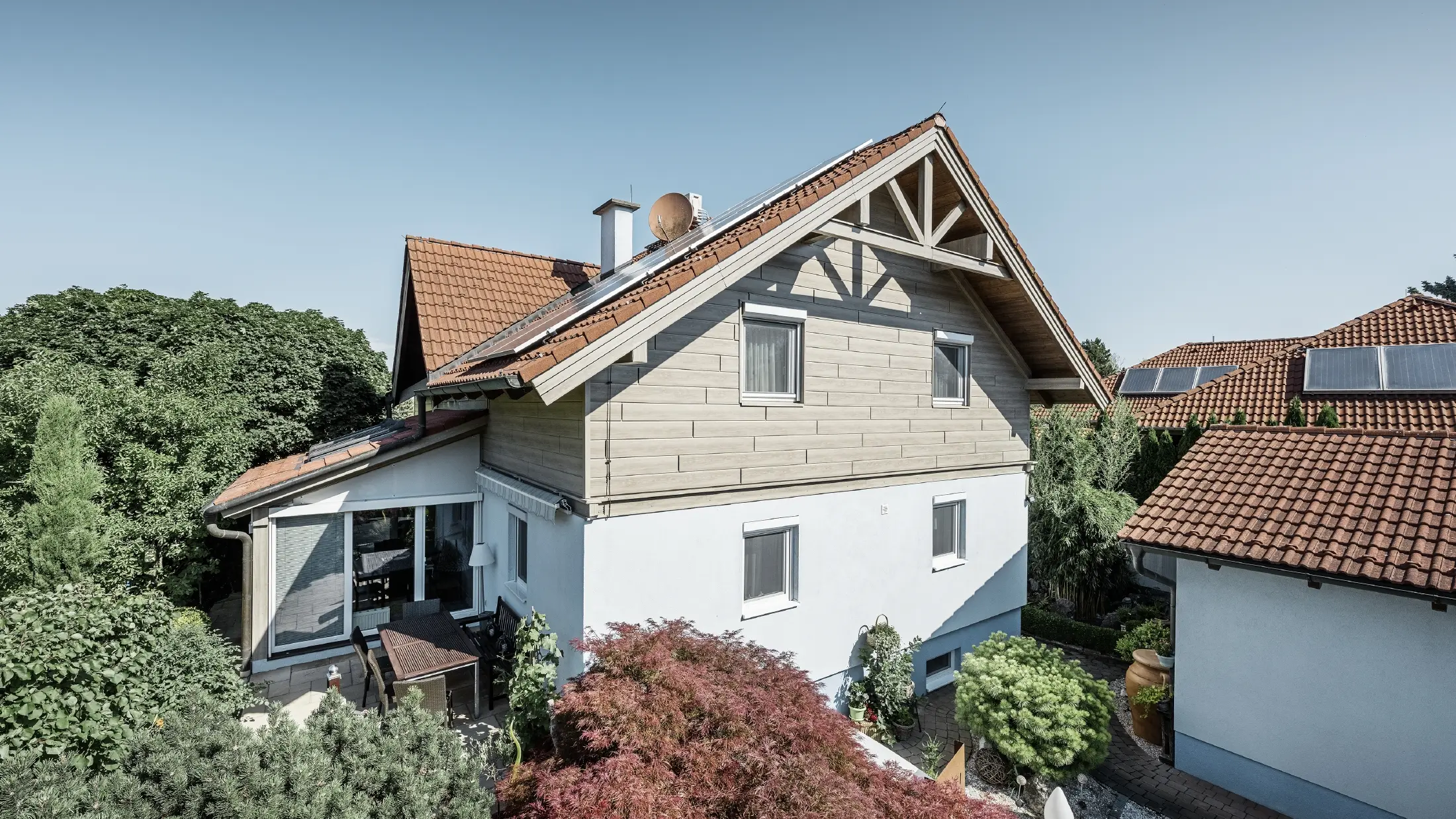 Casa unifamiliare con tetto a due falde e copertura rossa, abbinata a una facciata dal design moderno. Il frontone superiore è rivestito con pannelli per facciata in alluminio beige-grigio di PREFA, mentre il piano terra presenta una facciata intonacata di bianco. Sul tetto sono installati dei moduli fotovoltaici. La casa è circondata da un giardino verde con terrazza, mobili da giardino e piante rigogliose, ed è circondata da altre abitazioni con un'architettura del tetto simile.