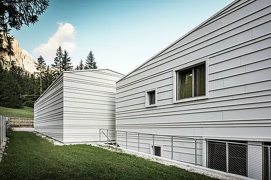 Il retro della caserma “Tempesti” a Corvara offre una prospettiva suggestiva: alberi del bosco, un cielo azzurro brillante e montagne illuminate dal sole. La facciata in alluminio chiaro con la sua struttura lineare mostra la raffinatezza tecnica di PREFALZ nel colore P.10 bianco PREFA. Le lamelle orizzontali in alluminio riflettono la luce, offrono una protezione ottimale dagli agenti atmosferici e si integrano armoniosamente nell'ambiente alpino. Un esempio eccellente di architettura militare moderna e durevole!