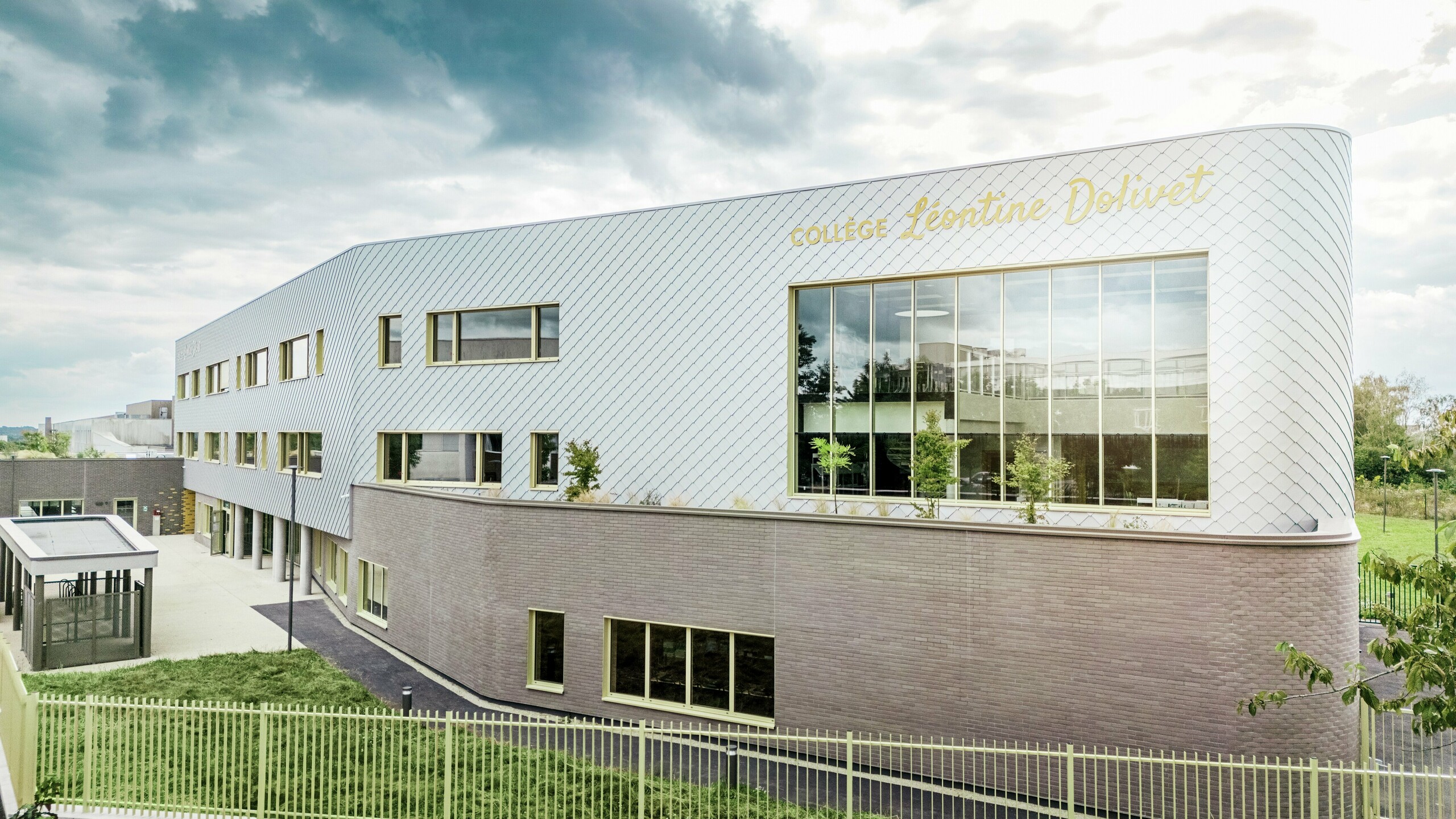 Seitenansicht des Collège Léontine Dolivet in Cesson-Sévigné mit moderner Aluminiumfassade aus PREFA Wandrauten in Silbermetallic. Die diagonale Struktur erzeugt einen dynamischen Look, während große Glasflächen und gelbe Fensterläden spannende Akzente setzen. Im Vordergrund sind Grünflächen und ein Zaun sichtbar, die das Gebäude einrahmen. Die langlebige PREFA Fassadenlösung verbindet Eleganz, Witterungsbeständigkeit und Nachhaltigkeit – ein architektonischer Eyecatcher mit klarer Linienführung und changierendem Metall.