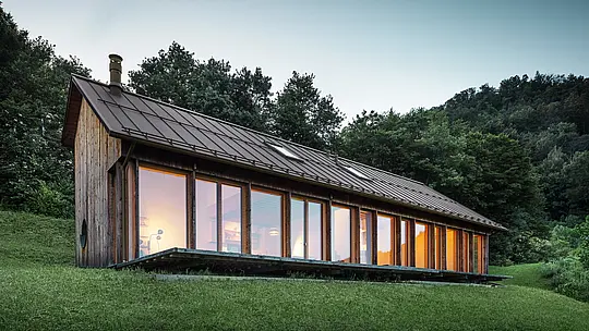 Abendliche Atmosphäre in einem modernen Holzhaus in Vinj mit durchgehender Glasfront und einem robusten PREFALZ-Aluminiumdach in der Farbe P.10 Braun. Die langlebige PREFA-Dachdeckung bietet zuverlässigen Schutz und fügt sich harmonisch in die natürliche Umgebung ein. Das Design vereint Wohnkomfort, Energieeffizienz und ästhetische Nachhaltigkeit.