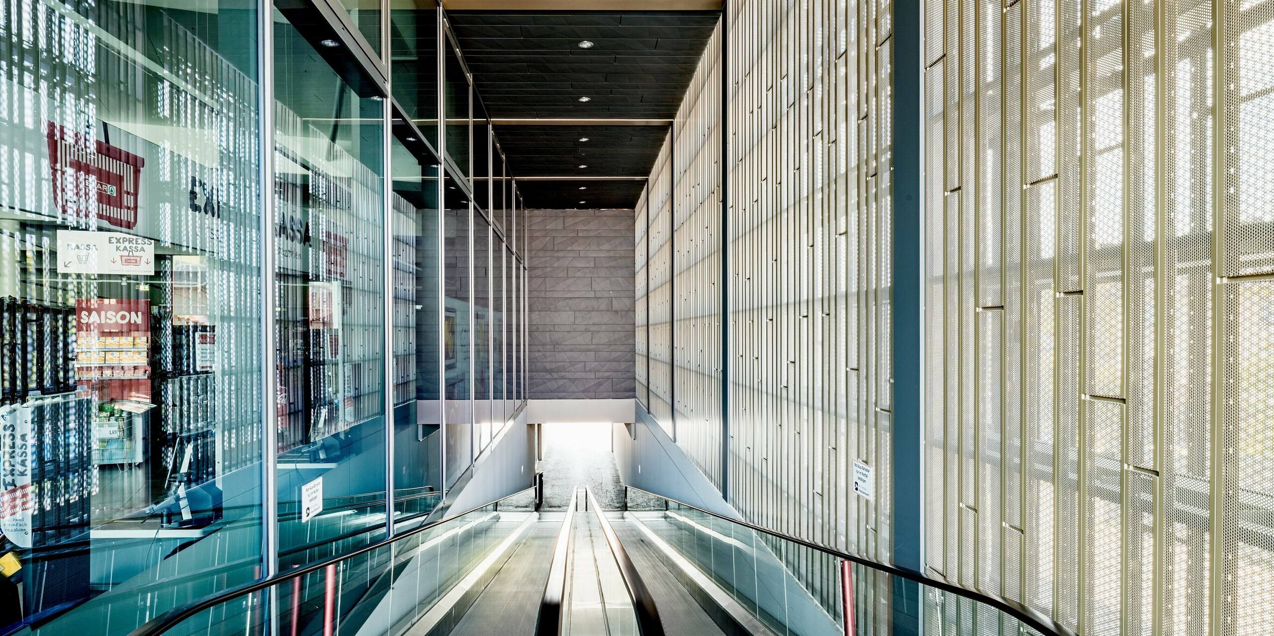 Das Bild zeigt den beeindruckenden Eingangsbereich des Einkaufszentrums in Rohrbach, der durch die Kombination moderner Materialien und funktionalem Design besticht. Im Vordergrund ist die Rolltreppe zu sehen, die die Besucher in die unteren Ebenen des Gebäudes führt. An den Wänden erkennt man die präzise angebrachten, perforierten Siding-Paneele in Anthrazit. Durch ihre leichte Textur und das spielerische Lichtspiel schaffen sie eine angenehme Atmosphäre. Diese Paneele sind nicht nur ästhetisch vorteilhaft, sondern sorgen auch für Belüftung und Lichtdurchlass und versorgen den Innenraum so optimal mit natürlichem Licht. Die großen Fensterflächen auf der linken Seite ermöglichen einen freien Blick nach außen und verstärken das Gefühl von Offenheit und Transparenz.