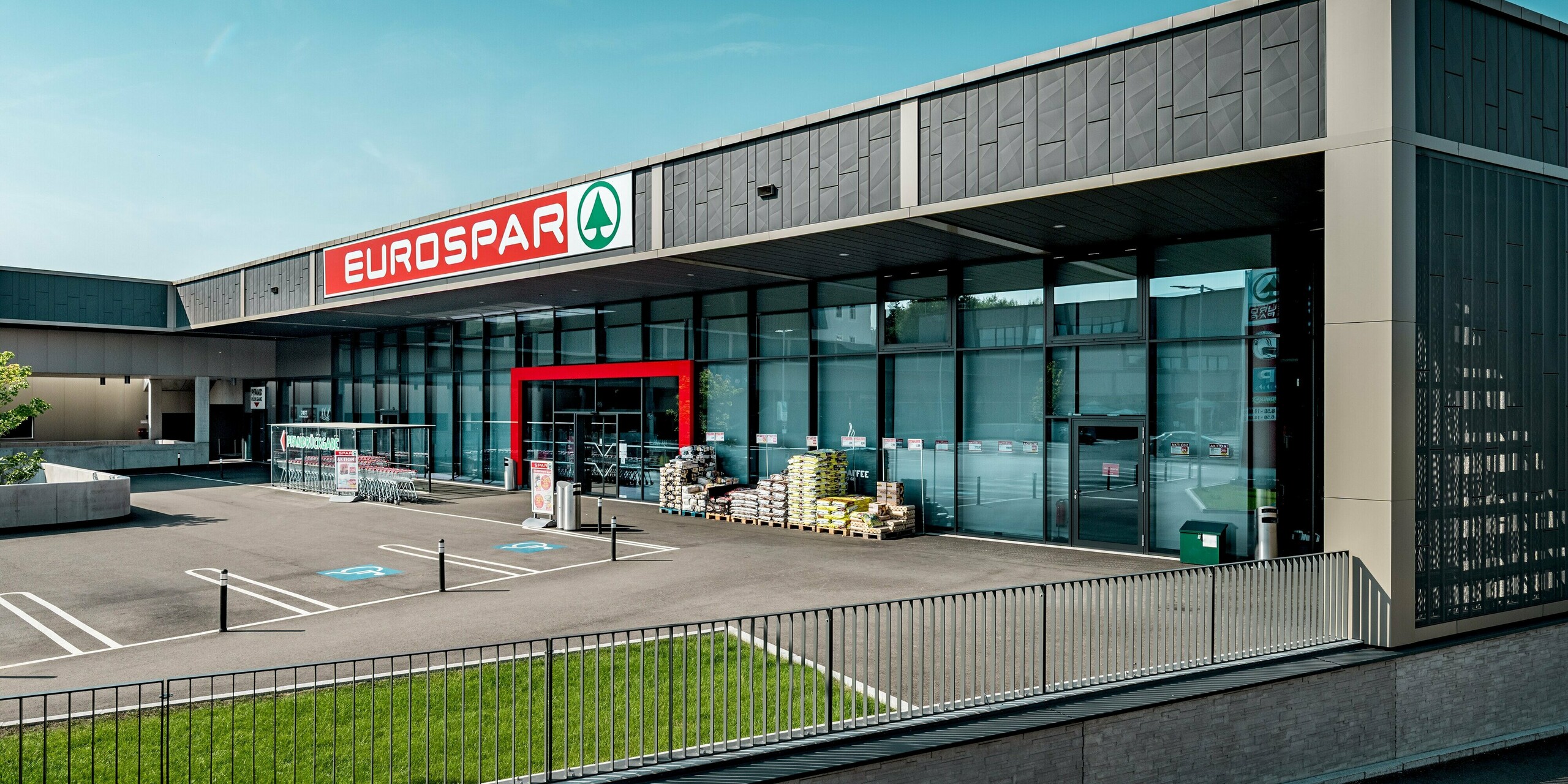 Der moderne Eurospar Supermarkt in Rohrbach wurde mit einer auffälligen Aluminiumfassade von PREFA verkleidet. Besonders bemerkenswert sind die anthrazitfarbenen Siding.X-Paneele, die dem Gebäude eine elegante und zeitgemäße Optik verleihen. Der markante Eingangsbereich ist durch eine typische, auffällige rote Umrahmung hervorgehoben, welche das Design zusätzlich betont. Die klar strukturierte Fassade mit dem Spiel zwischen Licht und Schatten verleiht dem Gebäude eine moderne und einladende Wirkung. Die Verwendung von PREFA Siding.X sorgt für eine stilvolle Optik und macht das Gebäude hoch witterungsbeständig und langfristig pflegeleicht.