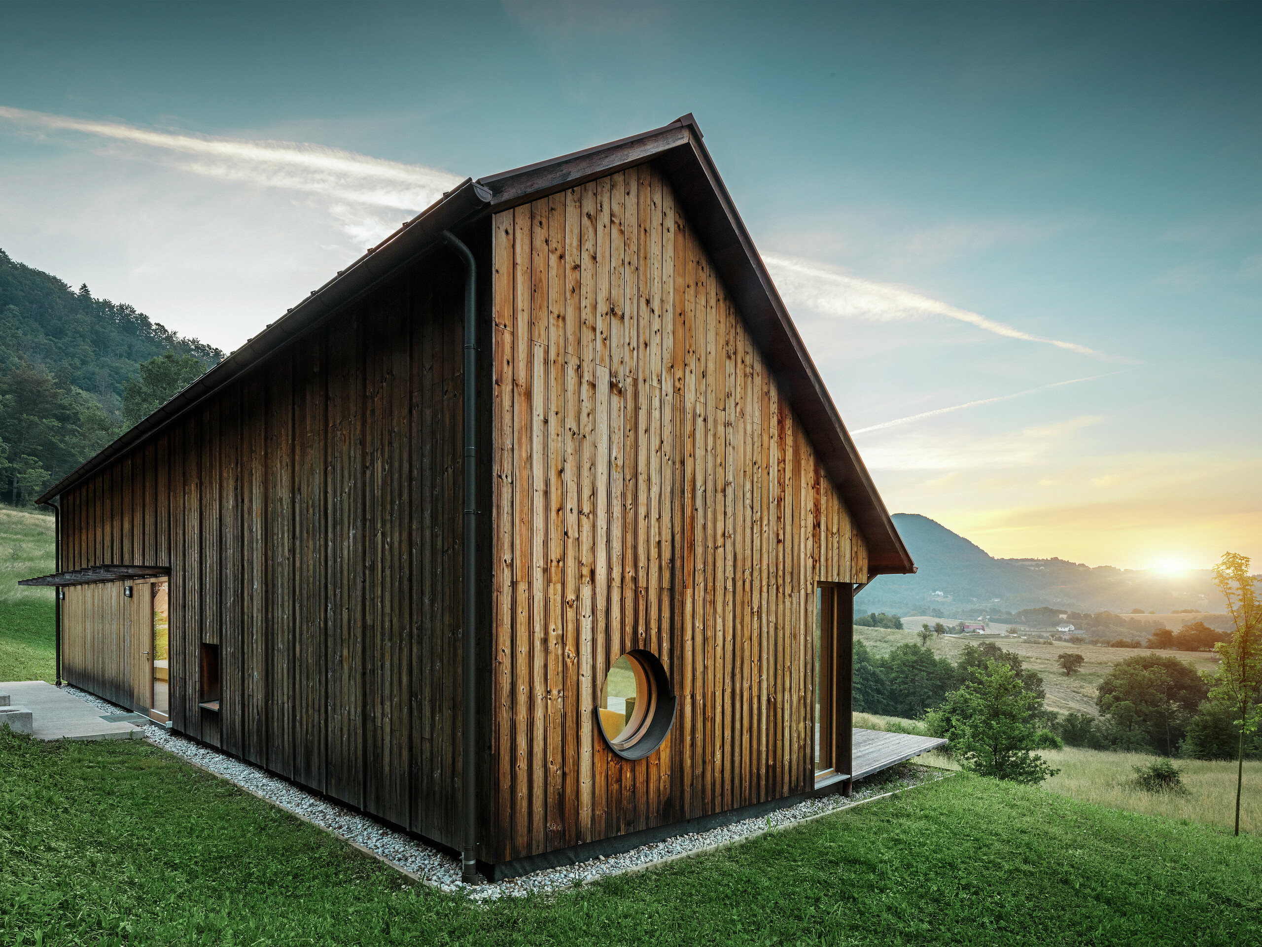 Questa suggestiva foto mostra una casa in legno a Vinje al tramonto con un robusto tetto in alluminio PREFALZ nel colore P.10 marrone. Il tetto in doppia aggraffatura PREFA, resistente agli agenti atmosferici, è durevole, richiede poca manutenzione e si integra perfettamente con l'ambiente naturale circostante. La combinazione di un linguaggio formale chiaro, la facciata in legno e l'apertura circolare delle finestre sottolineano il minimalismo architettonico. Il semplice tetto a falde è caratterizzato da una voluta asimmetria, che gli conferisce un tocco molto particolare.